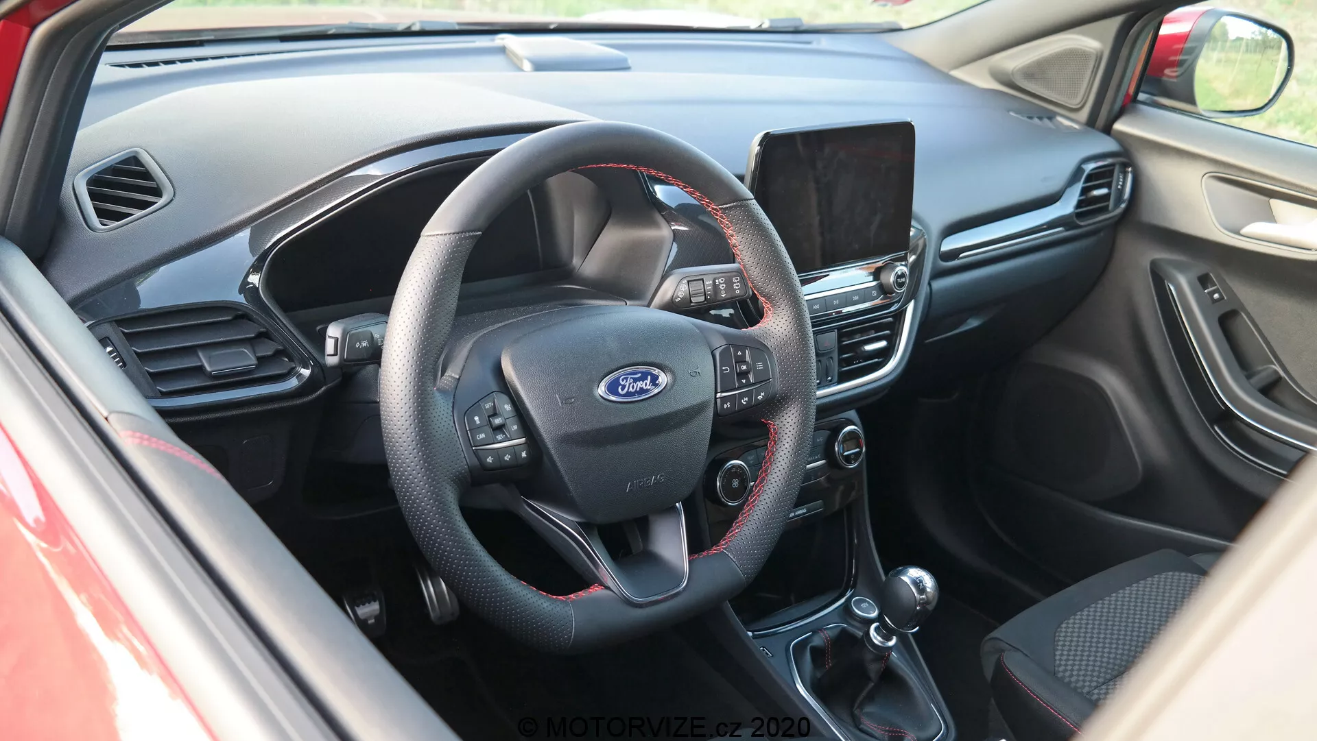 Interiörvy av Ford Puma 2020 cockpit från en något upphöjd vinkel, med fokus på mitt på instrumentbrädan. Sedd från förarsidan visar bilden en treekrad ratt med multifunktionsknappar för ljud, röstkommandon och farthållare. Fords logotyp är tydligt synlig i mitten. Bakom ratten syns en del av instrumentpanelen. I mitten av instrumentbrädan finns en infotainmentskärm som troligen erbjuder navigering, medieuppspelning och inställningar. Nedanför finns det manuella klimatkontrollsystemet med vred för temperaturjusteringar och knappar för luftkonditionering och avfrostning. Bilden avslöjar även automatväxelreglaget och handbromshandtaget. Interiören kombinerar mörk plast på övre delen av instrumentbrädan med svart tyg eller läder på de nedre delarna, vilket ger ett funktionellt designintryck med fokus på användbarhet.