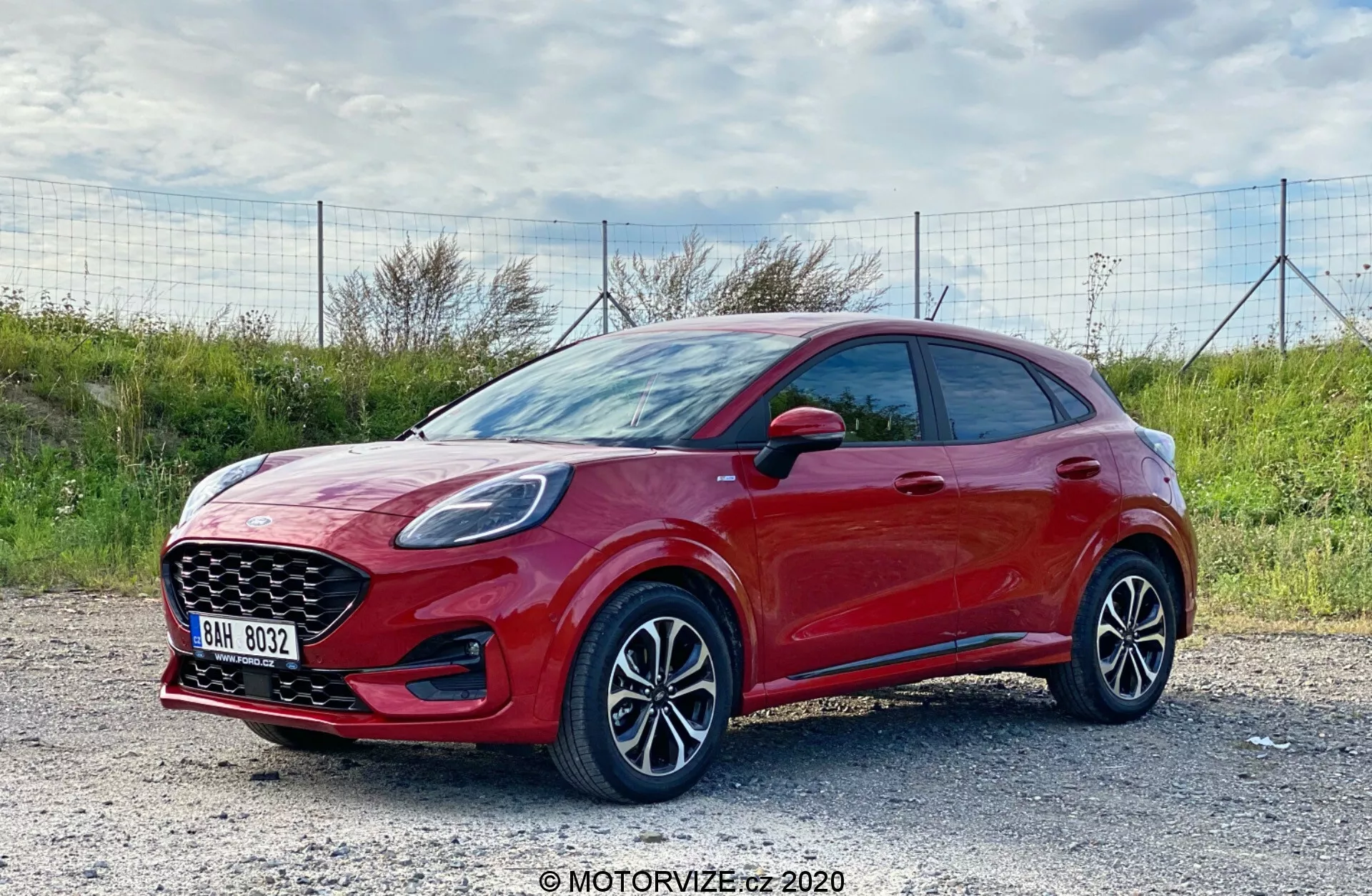 Sidoprofilbild av en modern hatchback Ford Puma 2020, fotad i dagsljus med en naturlig utomhusbakgrund. Det livfulla röda fordonet med en blank finish ses från en trekvartsvinkel framifrån, vilket tydligt visar dess sidoprofil och front. Bilen är parkerad på en grusväg. Bilden, tagen något ovanför hjulnivå, framhäver de mångekrade lättmetallfälgarna och den nedre delen av fordonet. Synliga är de eleganta strålkastarna, troligen med LED-teknik, en kraftig främre grill och sidospeglar med integrerade indikatorer. Bilen har karossfärgade dörrhandtag, inga synliga skyddande sidolister och en sportig hatchback-stil med en nedåtgående taklinje. Teknologiska funktioner kan inkludera avancerade förarassistanssystem och infotainment med anslutningsmöjligheter.