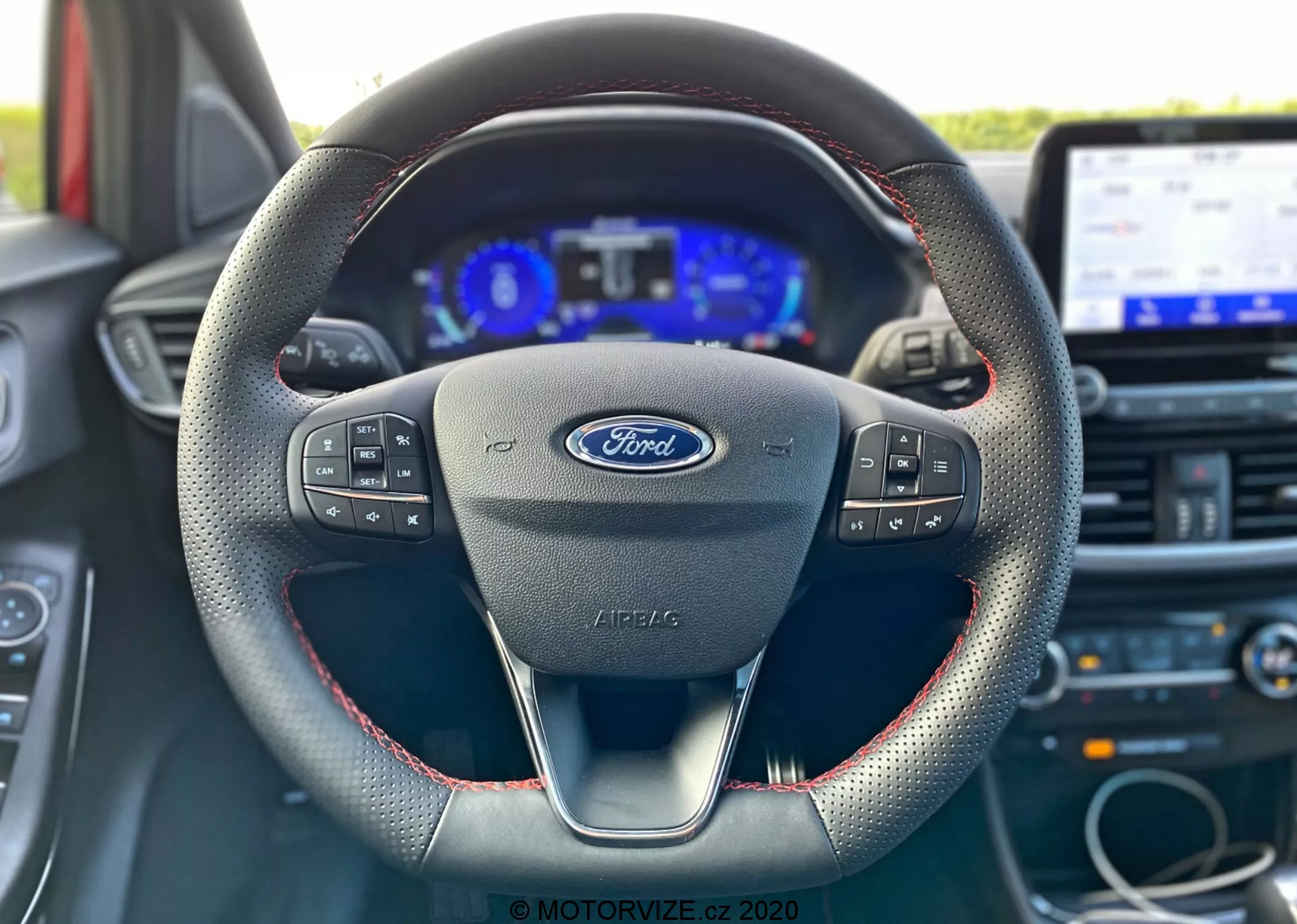 Interiörbild av en Ford Puma 2020 från förarsätets perspektiv, fokuserad på ratten med Ford-logotyp och multifunktionskontroller, en modern treekrad design med röda sömmar som antyder lädermaterial, en digital instrumentpanel flankerad av analoga mätare och en pekskärm för infotainmentsystemet i bakgrunden.