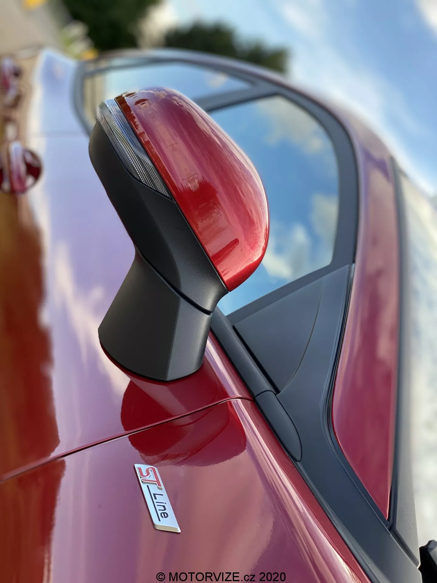 Närbild av förarsidans exteriöra backspegel på en röd bil, troligen en Audi med S Line-märke. Fotot taget från en låg vinkel fokuserar på backspegelenheten med ett grunt skärpedjup, vilket gör bakgrunden suddig. Spegeln är skarpt och detaljerat, visar integrerade blinkers, en strukturerad svart stöttning och en glansig, metallisk lack. Reflektioner av himlen och omgivningen indikerar att bilen är parkerad utomhus.