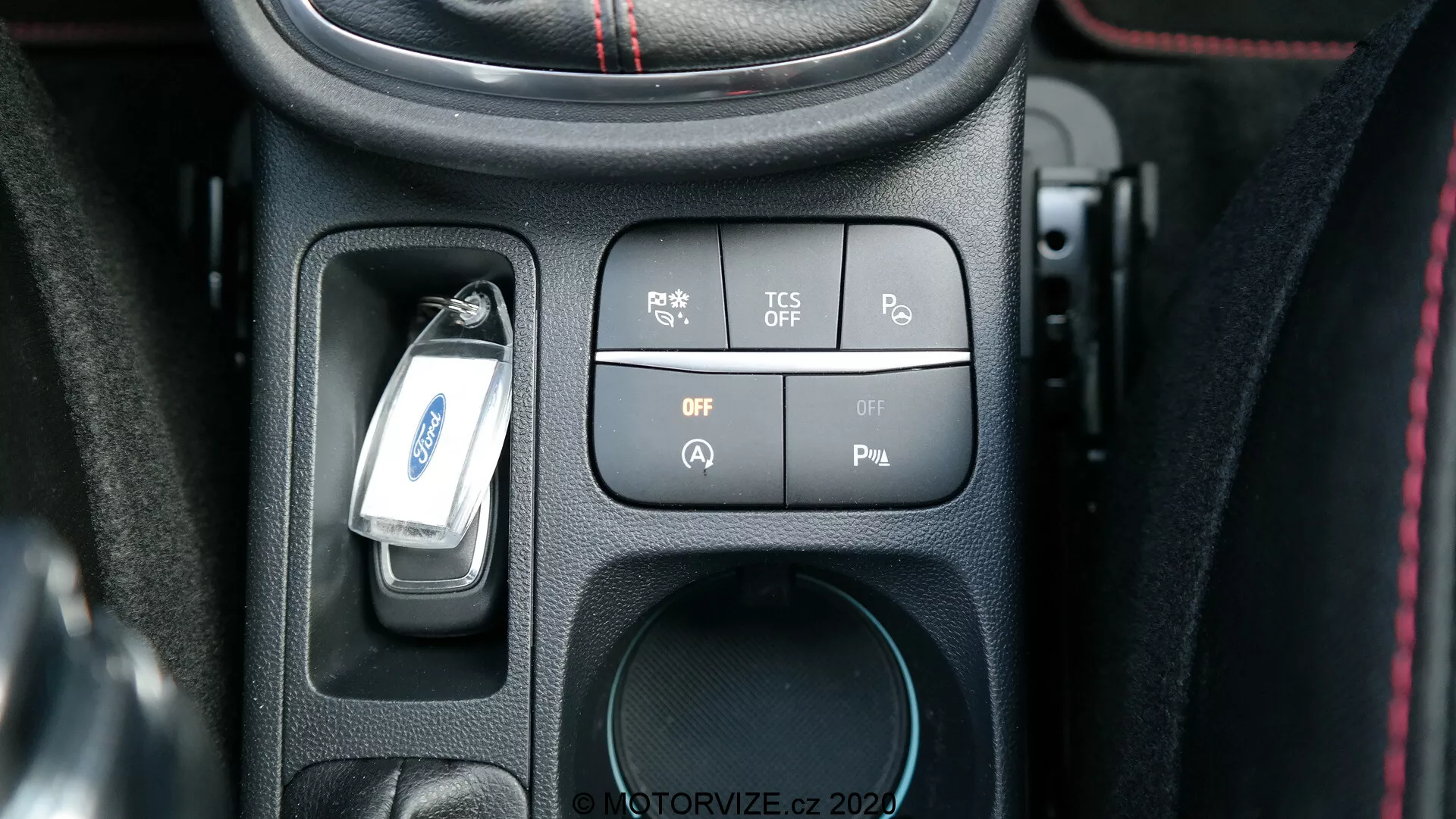 Närbild av den centrala konsolen på en Ford Puma 2020 från förarens perspektiv som visar en uppsättning högteknologiska knappar och strömbrytare för att kontrollera bilens funktioner. Konsolen inkluderar en parkeringssensor-knapp, en knapp för att avaktivera antispinnsystemet (TCS), två strömbrytare och en plats för ett 12V-uttag. En bilnyckel hänger över den centrala sektionen, och konsolen är accentuerad med en strukturerad svart plastinfattning och röda sömmar, vilket ger en premiumkänsla.