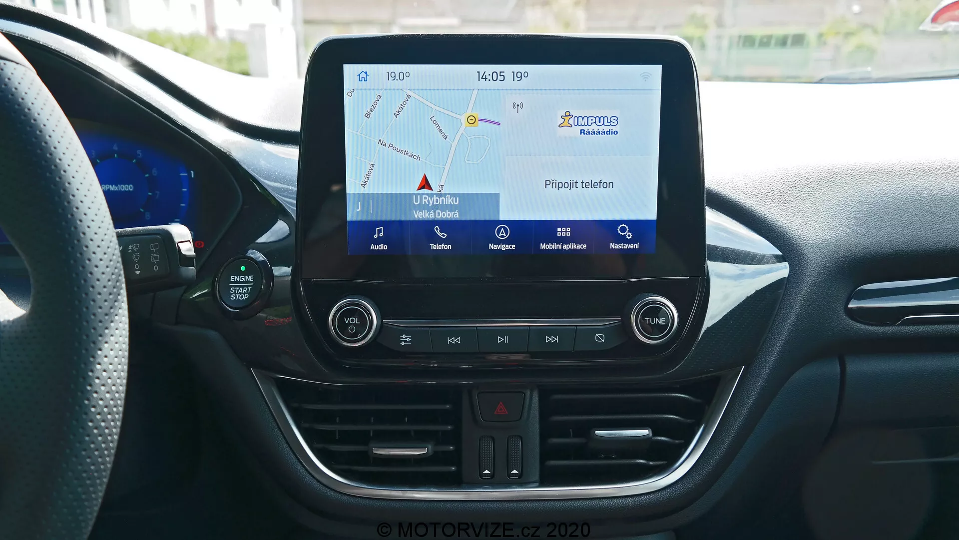 Mittkonsolen i Ford Puma 2020:s interiör från förarens eller passagerarens perspektiv, som visar infotainmentsystemet med pekskärm som visar navigationskartan, fysiska knappar för varningsblinkers, klimatkontrolljusteringar, CD-spelare, USB-port och 12V-uttag. Konsolen inkluderar även en digital termometer som visar 17°C och en klocka som visar 14:05, allt omgivna av moderna interiöra designelement med böjda linjer och luftkonditioneringsventiler.