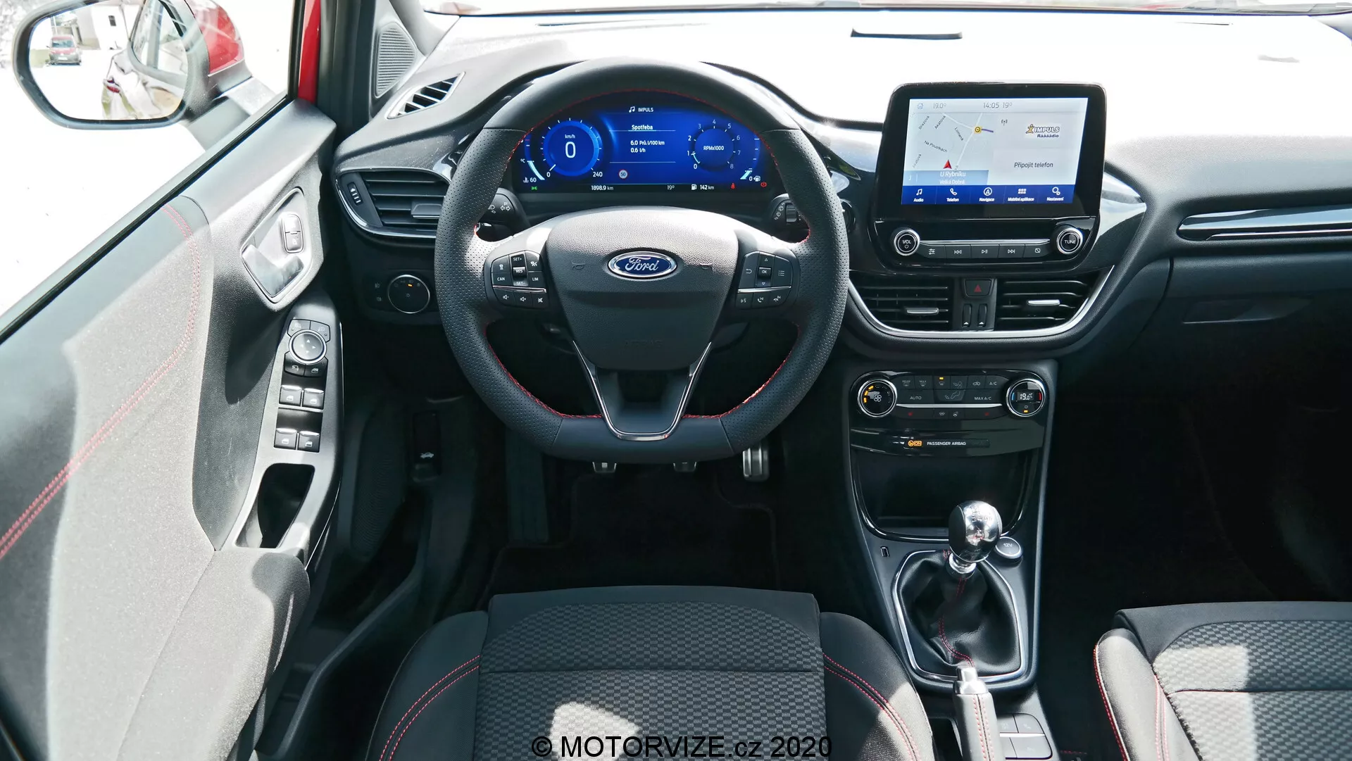 Interiörvy av Ford Puma 2020, tagen från den centrala bakre sittplatsen. Bilden visar en svart treekrad ratt med multifunktionskontroller och silverdetaljer, en digital instrumentbräda med blått tema, och en infotainment pekskärm som visar en karta, vilket antyder navigationsmöjligheter. Nedanför skärmen finns kontroller för luftkonditionering och ventilation med vrid- och tryckknappar för justering av temperatur och fläkthastighet. I mitten av tunneln finns växelspaken, vilket antyder en automatisk eller automatiserad manuell växellåda. Framsätena har tvåfärgade tygklädsel i svart med mönster för grepp och komfort, och en del av dörrens inre panel, inklusive ett silverhandtag och matchande detaljer, är synlig. Golvet är täckt av mattor som smälter in i färgschemat för interiören och det finns möjligen bra ambient belysning. Bilden är tagen från en något upphöjd vinkel som visar den främre interiören och tillgängliga teknologiska funktioner på ett omfattande sätt.