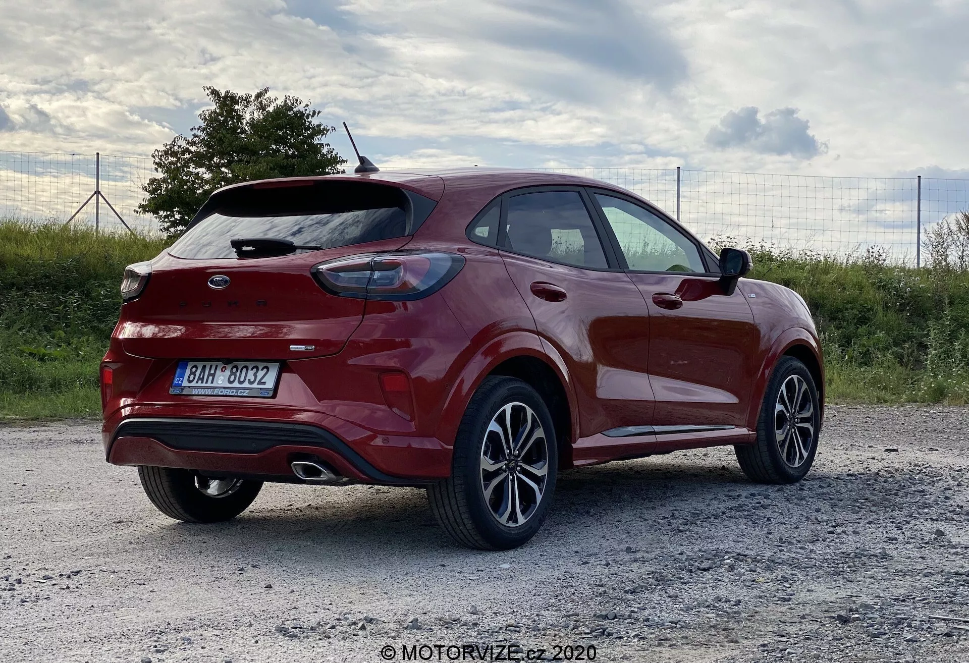 Den här bilden visar en modern Ford Puma crossover SUV från 2020, parkerad på en grusyta. Fotot är taget från en trekvarts bakifrån-vy och framhäver bilens sidoprofil och bakre delar, däribland den böjda bakluckan med integrerad spoiler, det framträdande emblemet, bakre vindrutetorkaren och de horisontella bakljusen. Den metalliska röda lacken glänser, vilket avslöjar en glansig finish kompletterad av lättmetallfälgar med multifliksdesign. Bilens kaross har karakteristiska linjer och kurvor för aerodynamik, med en stigande midjelinje och synliga takräcken. Belysningen på sen eftermiddag eller tidig kväll framhäver bilens färg och design. Potentiella teknologiska funktioner kan inkludera avancerade förarstödsystem (ADAS).