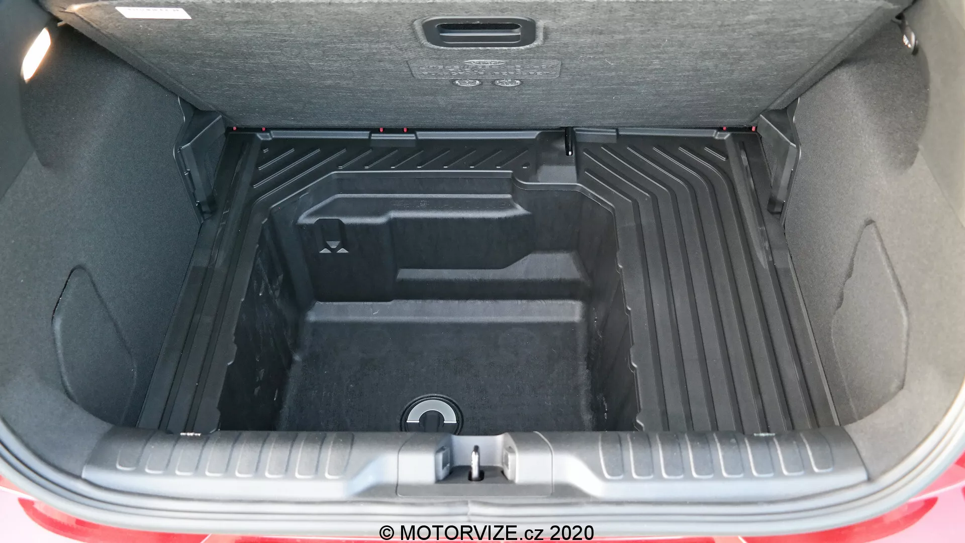 En detaljerad vy av det rymliga och mångsidiga bagageutrymmet i en Ford Puma 2020 med bakluckan öppen, som visar det tomma, rena och prydligt konturerade interiören utformat för organisering av bagage och last. Bagageutrymmets interiör är tillverkat av ett hållbart, lätt att rengöra material, med upphöjda och nedsänkta sektioner för optimerade förvaringslösningar, inklusive ett centralt nedsänkt handtag för att komma åt ett nedre fack eller ett reservhjulsutrymme. Den bakre stötfångaren och en remsa av bilens röda exteriörfärg är delvis synliga, tillsammans med det isolerade bagagelucklocket utrustat med ett draghandtag för enkel stängning.