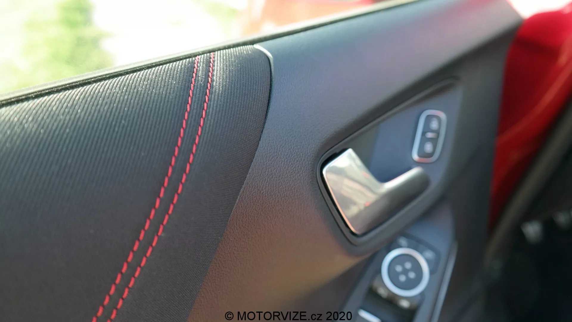 Närbild av 2020 Ford Pumas interiöra dörrpanel ur en passagerares eller förares perspektiv inuti fordonet. Bilden fokuserar på handtagsområdet, framhäver den texturerade insatsen med röd kontrastsöm, metalliskt dörrhandtag och en del av kontrollpanelen med strömbrytaren för elfönstret. Det grunda skärpedjupet gör bakgrunden suddig, betonar de sportiga och lyxiga designelementen i naturligt dagsljus.