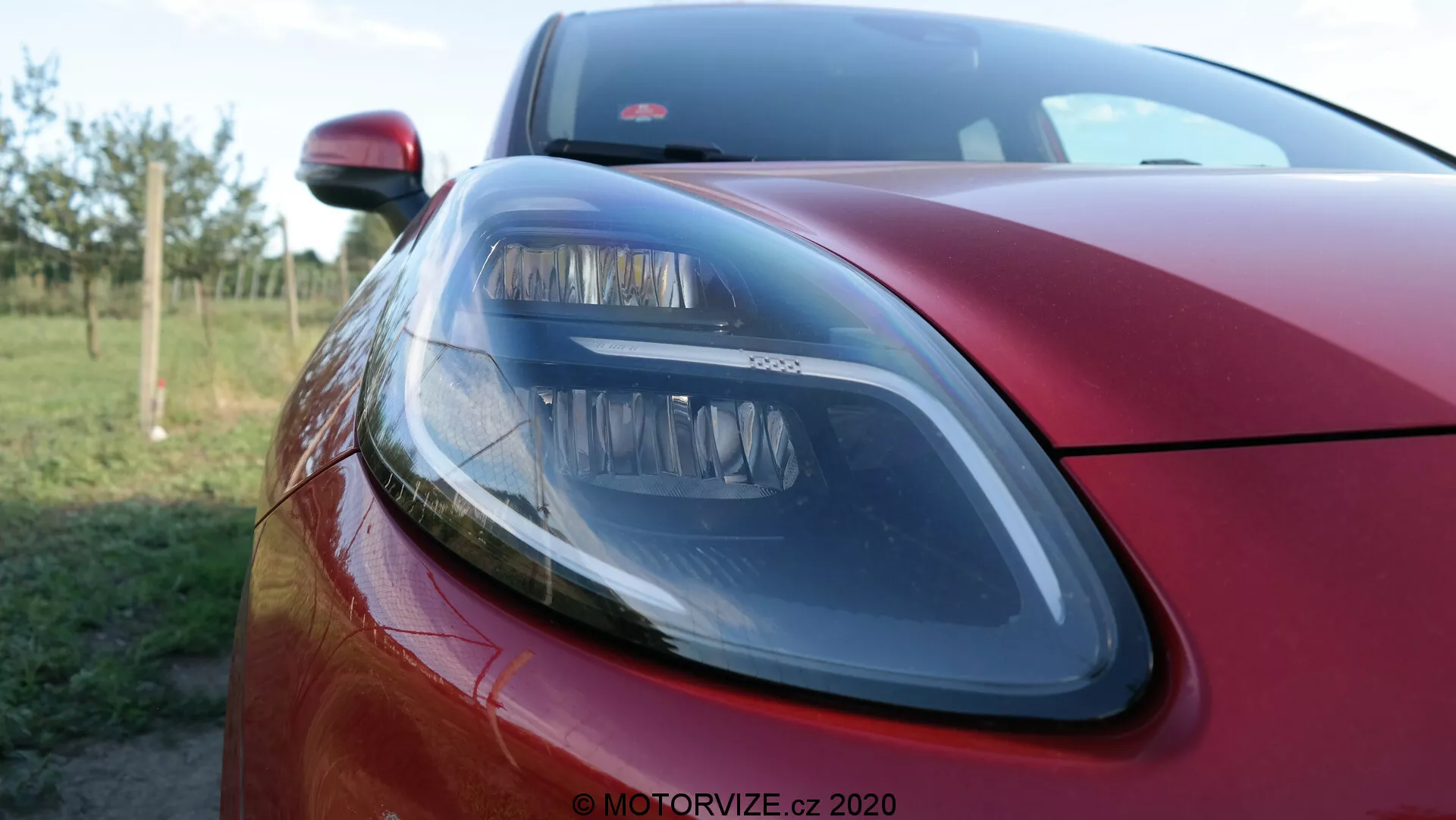 Närbild av den vänstra främre delen på en röd metallisk 2020 Ford Puma, som visar upp en modern strålkastare med eleganta designelement, en del av motorhuven och en glimt av frontgrillen med ett grunt skärpedjup som suddar ut bakgrunden, vilket framhäver den premium exteriöra finishen och belysningsteknologin.