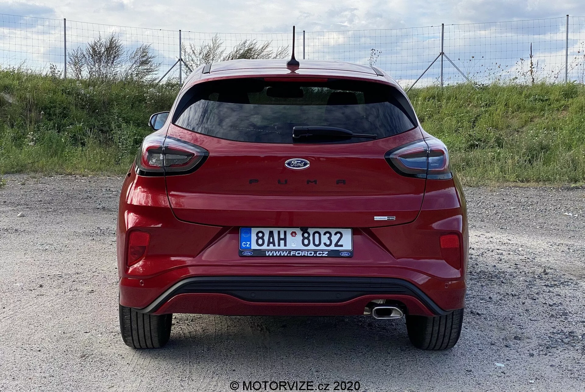 Bakifrån vy av en röd Ford Puma från 2020 hatchback på en grusig yta, fotograferad ur en nivå och centrerad vinkel. Funktioner inkluderar en bakspoiler, bakrutetorkare, högt monterad bromsljus, dubbla avgasrör, reflekterande ytor, parkeringssensorer, backkamera och tydligt visat Ford-emblem och modellnamn. Inget dragutrustning eller takräcke synligt. Foto av MOTORVIZE.cz.
