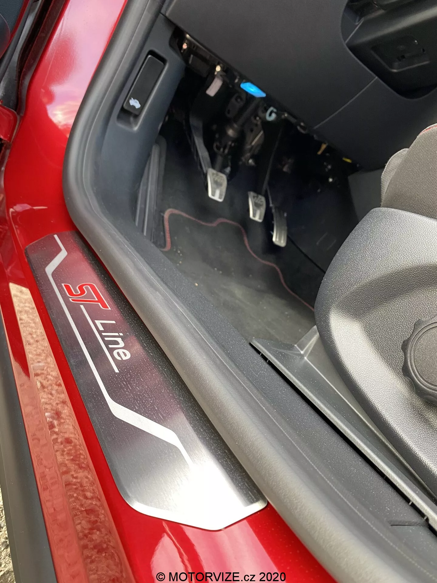 Närbild av dörrtröskeln på en Audi med 'S Line'-badge i silver och röd på svart bakgrund. Bilden tagen ovanifrån och något inåt fångar det nedre dörrkarmen, den inre kanten och en del av förarens fotutrymme med synliga broms- och gaspedaler. Naturligt dagsljus belyser scenen, vilket lyfter fram den röda exteriörfärgen och interiörkomponenterna nära instrumentpanelen.