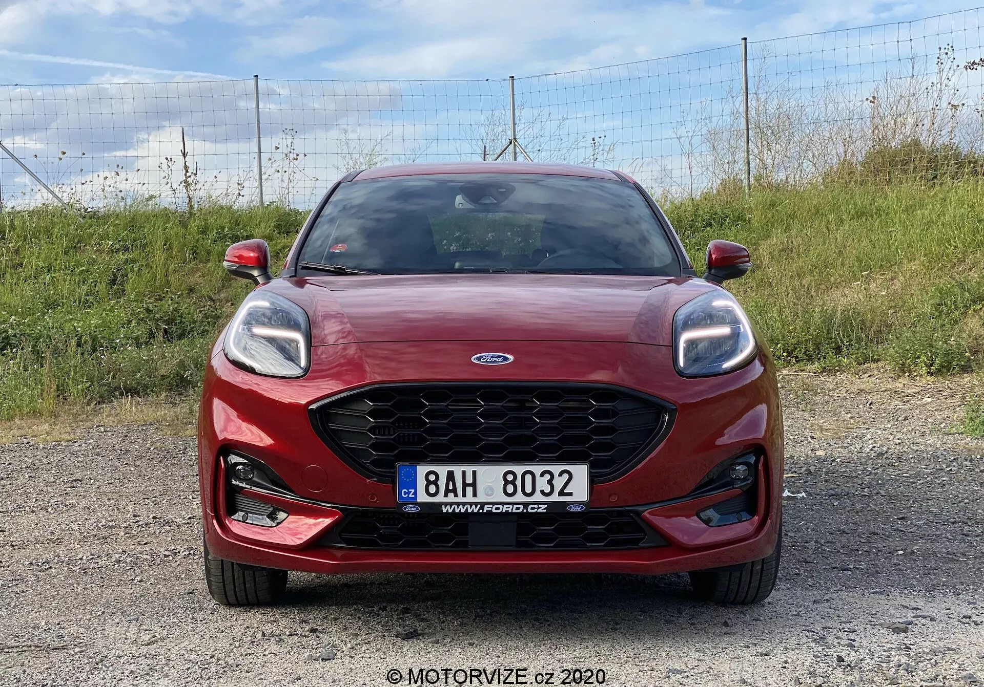 Röd Ford Puma 2020 fotograferad framifrån på en grusbelagd yta, visar den karakteristiska Ford-grillen med centrerad logotyp, eleganta strålkastare med LED-varselljus, integrerade dimljus och centrerad registreringsskylt. Perfekt för fordonets frontdesign, varumärkesidentifiering och moderna bilbelysningssystem.