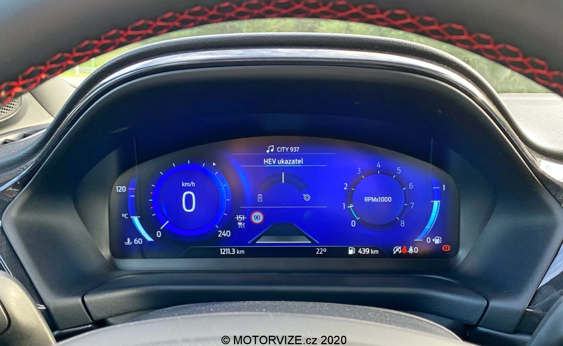 Närbild av Ford Puma 2020:s moderna digitala instrumentbräda från förarens perspektiv. Den digitala displayen visar en hastighetsmätare med ekovänliga hastighetsintervall till vänster, en varvräknare i mitten, hybriddrivmedelsmätare till höger och ytterligare information som körsträcka, resväg, räckvidd och utomhustemperatur längst ner. Ett meddelande högst upp i mitten säger 'HEV úzkazník!', vilket indikerar hybridläge för elfordon. Den eleganta och teknikinriktade designen i nyanser av blått, vitt och rött framhäver de avancerade förarinformationssystemen och hybridtekniken i fordonet.