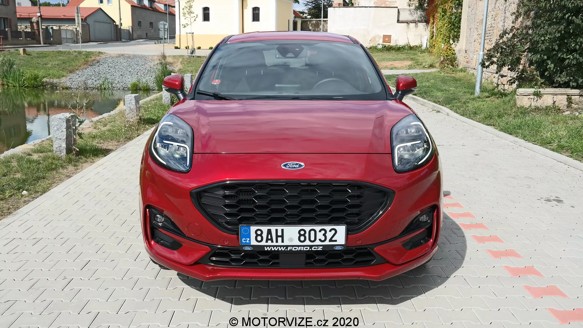Frontal vy av en röd 2020 Ford Puma som visar ett hexagonalt galler med svart nät, moderna svepande strålkastare, sportig frontstötfångare med breda luftintag och central registreringsskylt. De dynamiska konturlinjerna på motorhuven förstärker fordonets estetik. I bakgrunden syns ett bostadsområde.