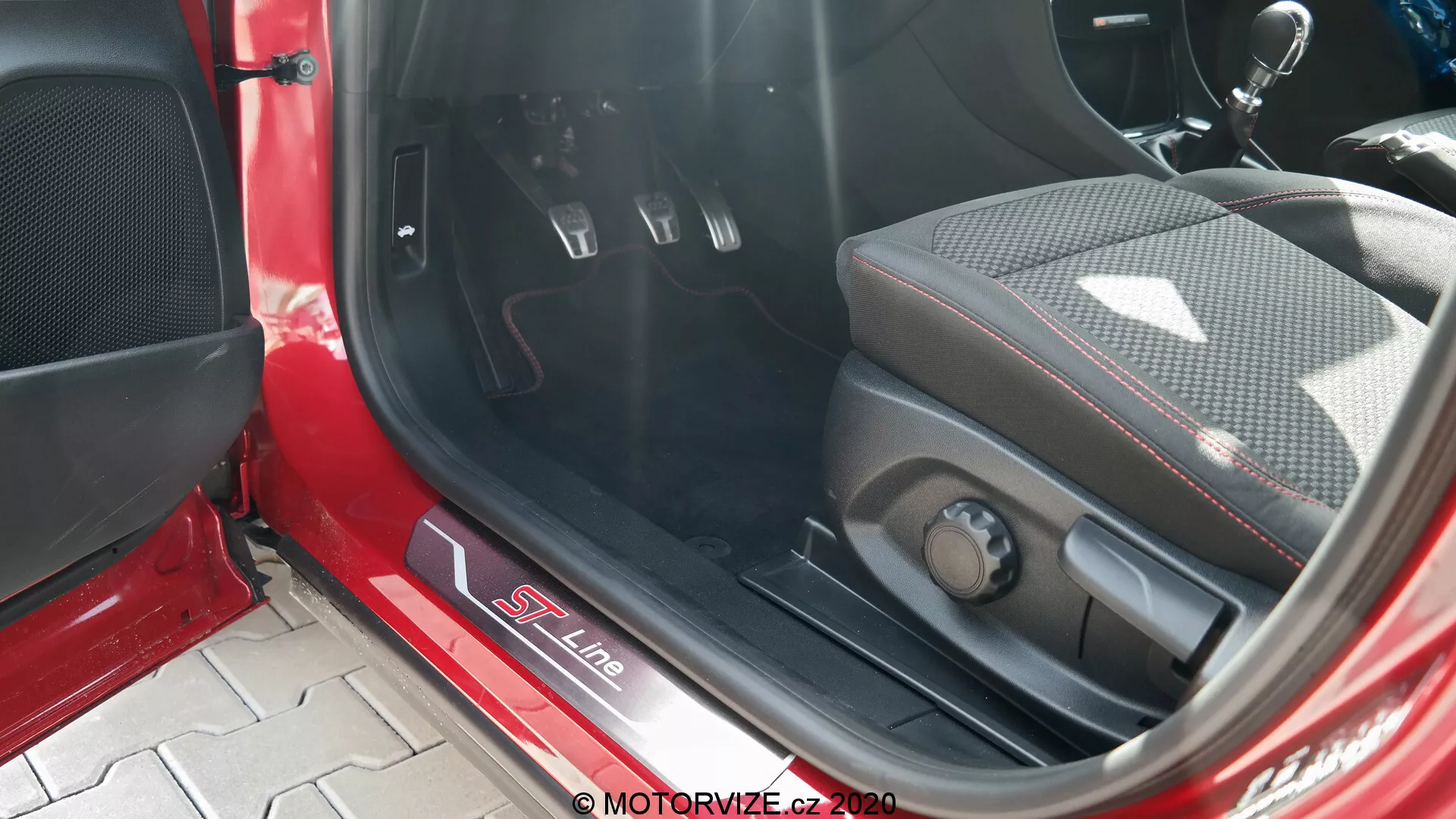 Närbild av interiören i en Ford Puma 2020, med fokus på S Line-märkt metallisk tröskelplåt, sportigt säte i en blandning av läder och tyg med sidostöd, elektroniska sätesjusteringskontroller, metallpedaler med gummiinlägg för manuell växellåda, mattbelagd golvmatta, röd och svart interiörpanel med högtalargaller och direkt solljus som lyser upp interiören.