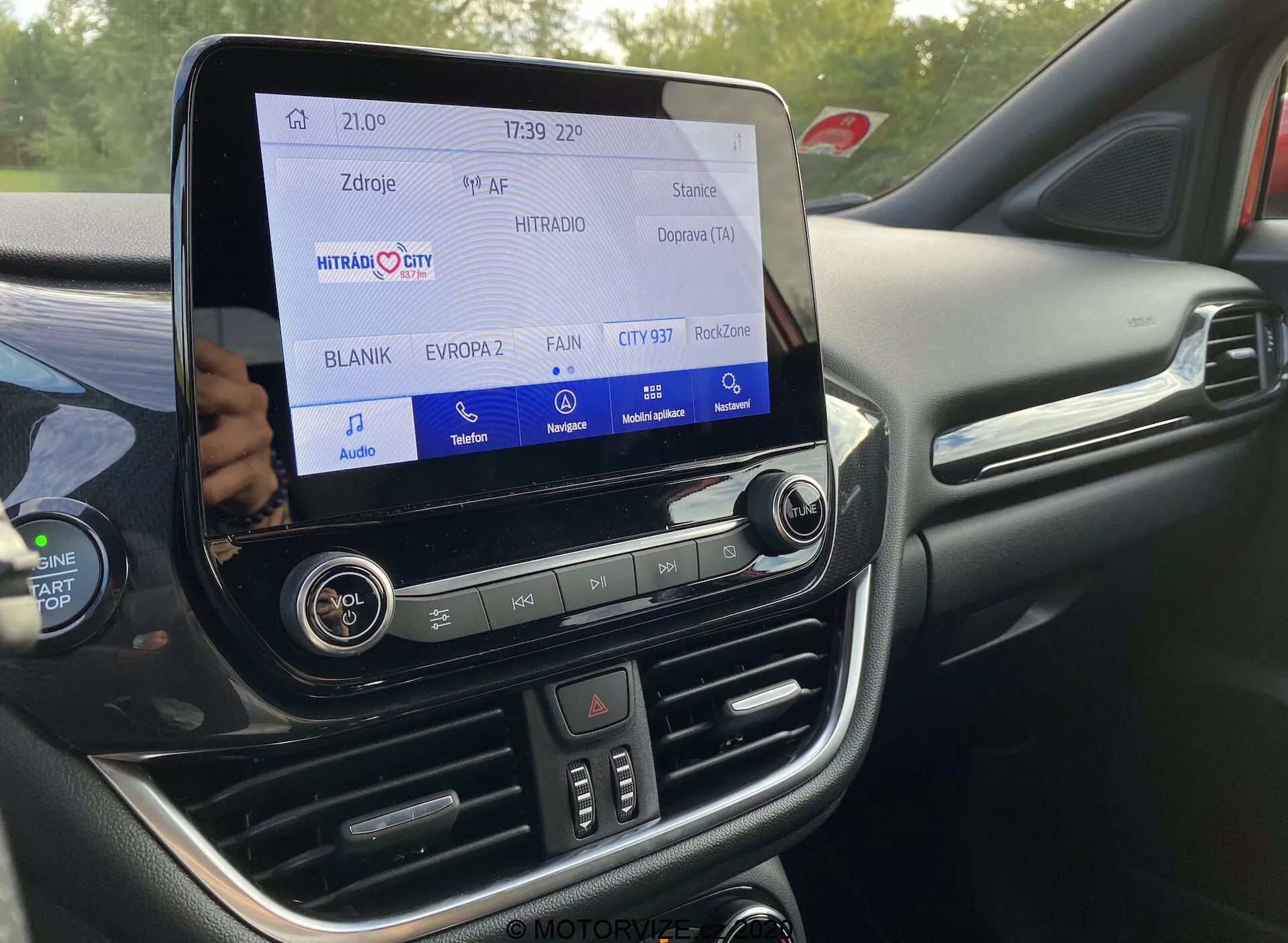 Närbild av interiören i Ford Puma 2020, med fokus på det avancerade infotainmentsystemet med en pekskärm som visar radio/media-gränssnittet, tiden 21:09 och 4G/LTE-anslutning. Fysiska knappar och reglage för manuell kontroll, tvåzons klimatanläggning med oberoende temperaturinställningar, och en premium svart instrumentpanel med sömmar och mjuka material är synliga. Rattknappar och en metallisk dörrhandtag antyder bilens högkvalitativa design.