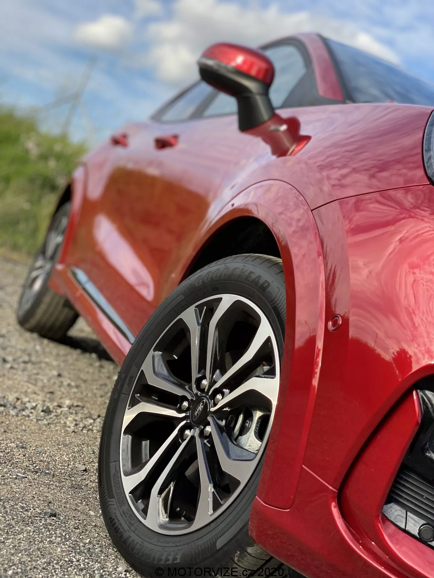 Närbild av en röd Ford Puma 2020 sportbil, som fokuserar på bakhjulet och fjärdedelspanelen. Den låga vinkeln, nära marknivån, framhäver den sofistikerade flerspakiga hjuldesignen, lågprofildäcket och den glänsande lacken. Bilden fångar invecklade detaljer av fälgdesignen och mönstret på slitbanan, med ett litet skärpedjup som håller hjulet skarpt i fokus medan bakgrunden är suddig. En liten luftintag syns bakom hjulet på fjärdedelspanelen, vilket betonar fordonets prestanda och aerodynamiska funktioner.