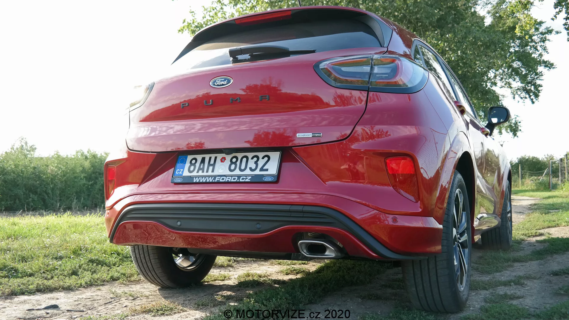 Bild av en röd Ford Puma 2020 fotograferad från en bakre trekvarts vinkel, som visar dess moderna design som hatchback/crossover-SUV. Bakluckan har en integrerad spoilervinge, omfattande horisontella LED-bakljus och synliga märken. Bakre stötfångaren inkluderar integrerade reflexer och ett krombelagt avgasrör. En del av det bakre legeringshjulet med flerspeksdesign är synligt. Bilen är parkerad utomhus i en naturlig miljö, vilket tyder på ett förorts- eller landsbygdsområde.