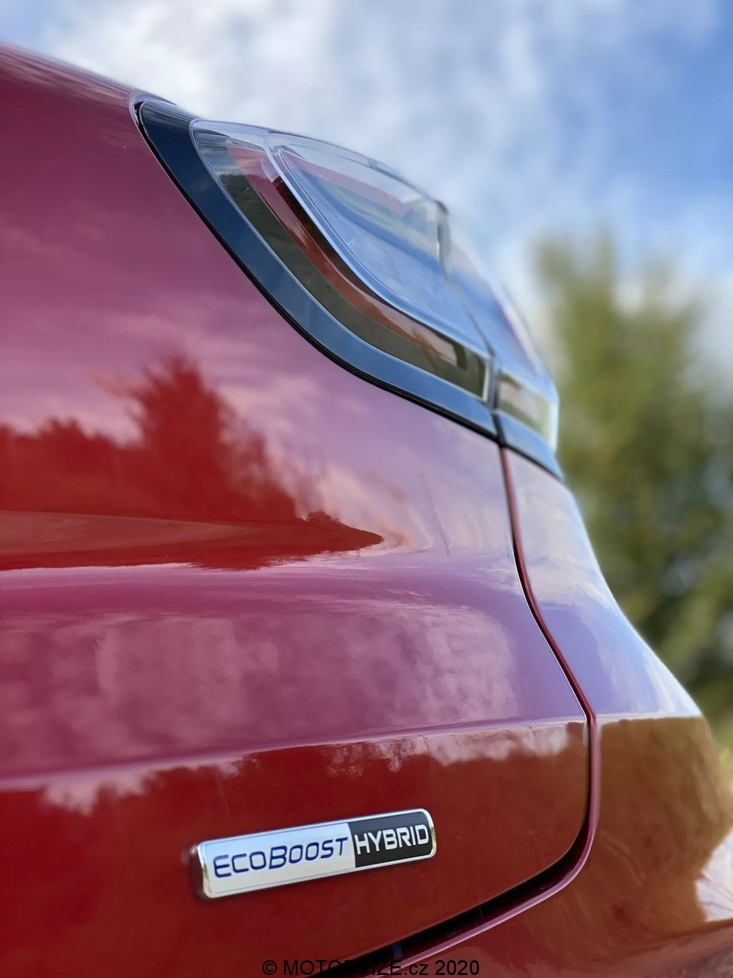 Närbild av bakre högra sidan på en röd Ford Puma 2020, med fokus på LED-baklyktan och bakre skärmområde från en låg vinkel med EcoBoost Hybrid-emblemet synligt, som reflekterar en blank finish och elegant design mot en bakgrund av himmel och lövverk.