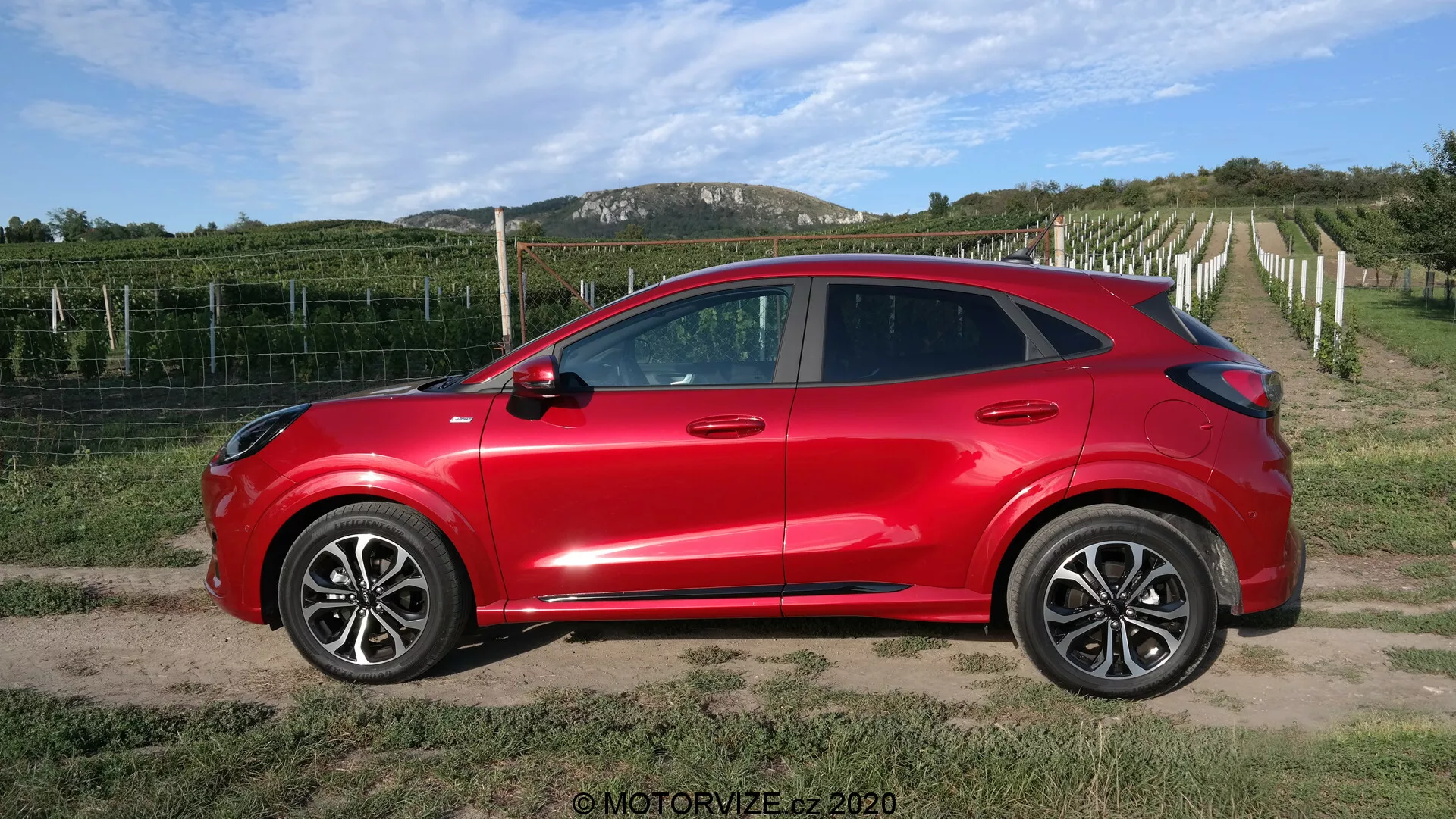 Sidprofilvy av en ljusröd 2020 Ford Puma kompakt crossover SUV med fyra dörrar, stora legeringshjul, sidospeglar i karossfärg med integrerade blinkers, tonade rutor och strömlinjeformade dörrhandtag i karossfärg. Fordonet med sin moderna aerodynamiska design och sluttande taklinje är placerat på en grus- eller grusväg med en vingård och klar himmel i bakgrunden, vilket ger en lantlig miljö.