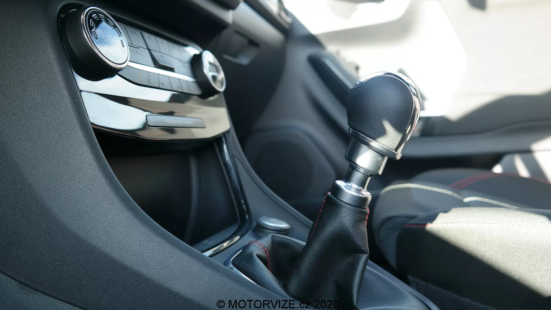 Interiör av en Ford Puma 2020-bil, med fokus på mittkonsolen från passagerarens perspektiv. Bilden framhäver en sexväxlad manuell växelspak i mörk plast med glänsande pianolack runtom basen. I bakgrunden syns den delvis suddiga ratten med ljud- och farthållarkontroller samt en ventilationsgaller på instrumentpanelen. Scenen är belyst av naturligt dagsljus.