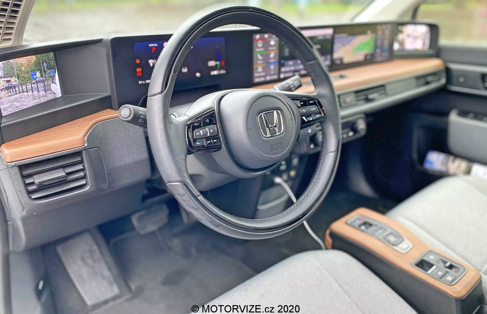 Interiör av Honda E 2020 som visar förarsidan med fokus på ratten och den moderna instrumentbrädan. Närbild framhäver Honda-logotypen på ratten, som har monterade kontroller för multimedia och farthållare. De förlängda, oavbrutna skärmarna fungerar som digitalt instrumentkluster som visar hastighet och infotainmentsystem med navigationskarta. Synlig träpanel ger en touch av lyx, och de ljusa tygklädda sätena indikerar både komfort och stil. Avancerade funktioner som pekskärmar, startknapp och nyckellös tändning understryker den teknologiskt utrustade kupén.