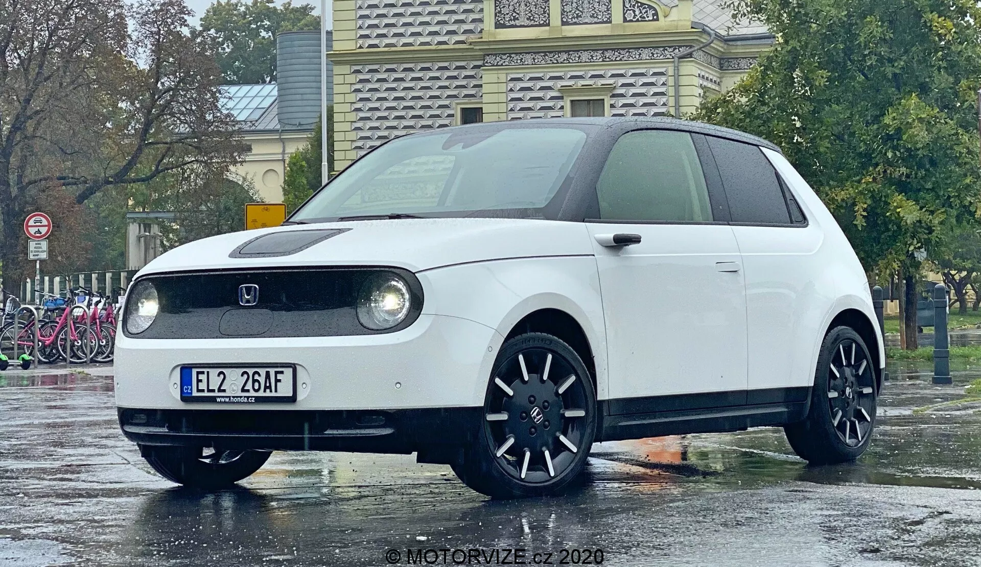 2020 Honda e fotograferad från en låg vinkel som betonar dess front och delvis profil från sidan. Bilden framhäver runda LED-strålkastare i rektangulära höljen, ett stängt frontgaller typiskt för elfordon, en centralt placerad tillverkarens emblem, mörka flerspokes fälgar och en minimalistisk retro-modern design. Den kompakta hatchbacken står på en våt väg vilket speglar en regnig miljö och framhäver bilens rena linjer och färg.