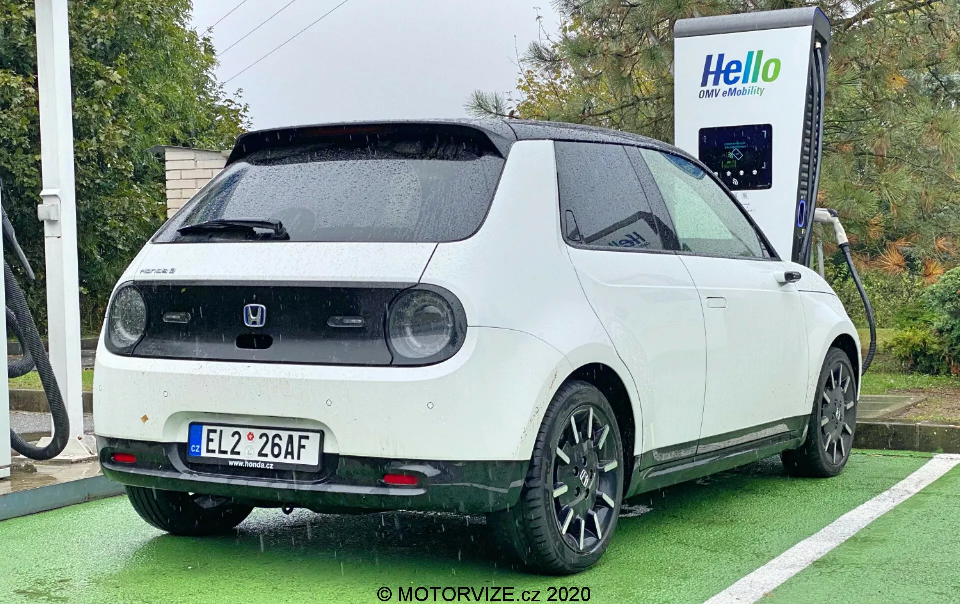 En 2020 Honda e elektrisk fordon fotograferad från en bakre trekvartsvinkel, vilket visar högra bakre delen, delvis sidovy och baklucka. Bilen, med en tvåfärgad färgschema med vit kaross och svart tak, är parkerad vid en laddningsstation med laddkabeln ansluten till porten på bakre sidan. Fordonets design inkluderar runda baklyktor, en stor bakre vindrutetorkare, parkeringssensorer, en takspoiler och svarta femekrade lättmetallfälgar. Bilden framhäver även den fyrkantiga, retro-moderna estetiken av bilen, tagen under dagtid i molnigt eller regnigt väder, med bilen parkerad i en särskilt anvisad laddstation för elbilar, markerad med grön färg.