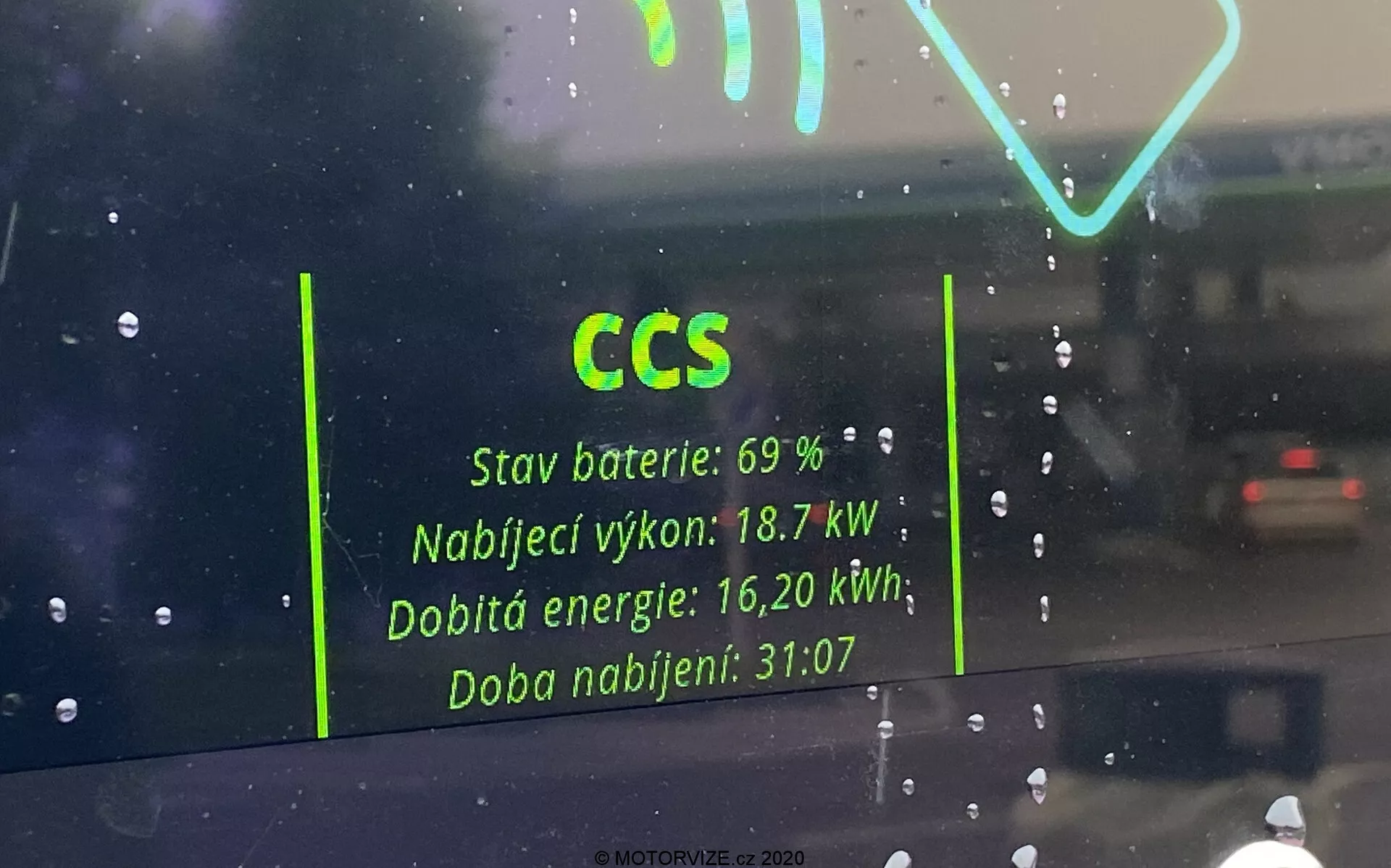 Interiörvy genom vindrutan på ett elfordon, med fokus på reflektionen av en laddstationsgränssnitt på glaset. Nyckeldetaljer inkluderar 'CCS' för Combined Charging System, 'Batteristatus: 69 %,' 'Laddeffekt: 18,7 kW,' 'Återuppladdad energi: 16,20 kWh,' och 'Laddningstid: 31:07.' Reflektionen framträder spöklik mot en regnig bakgrund med en annan bil synlig på avstånd.