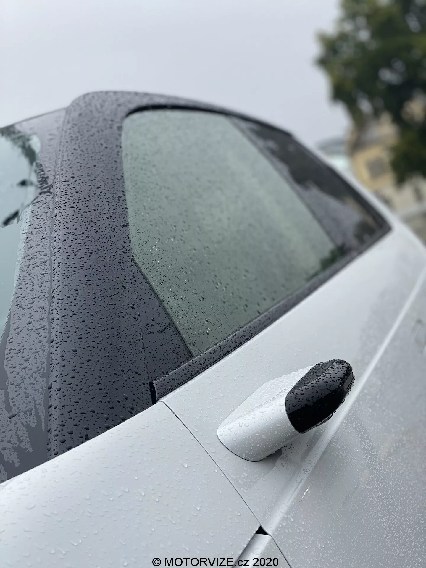 Närbild av bakre delen av Honda e 2020, som framhäver designen på det bakre passagerarsidans fönster med vattendroppar och den rena, ljusa bakdörren med ett svart utdragbart dörrhandtag, vilket betonar avancerad lackbeläggning och nyckelfri entré.