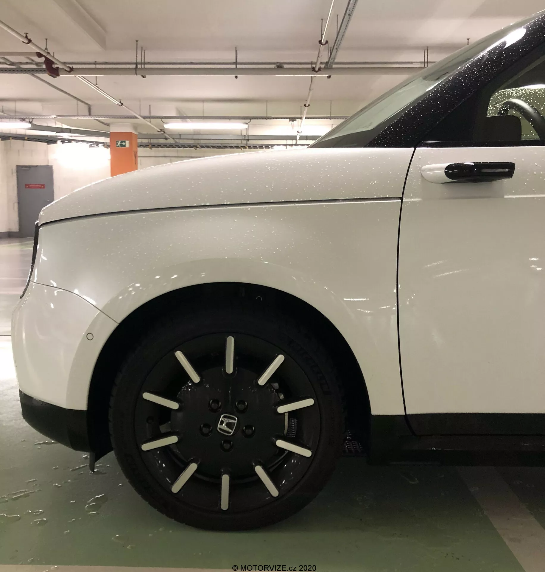 Närbild från sidan av framhjulet och kvartspanelen på den vita Honda E 2020, som framhäver det tio-ekrade lättmetallfälgen och den rena, aerodynamiska karosserin. Bilden, som sannolikt tagits i ett inomhusparkeringsgarage, betonar fordonets välskötta skick och robusta design, med synliga våta ytor som indikerar nylig exponering för vatten.