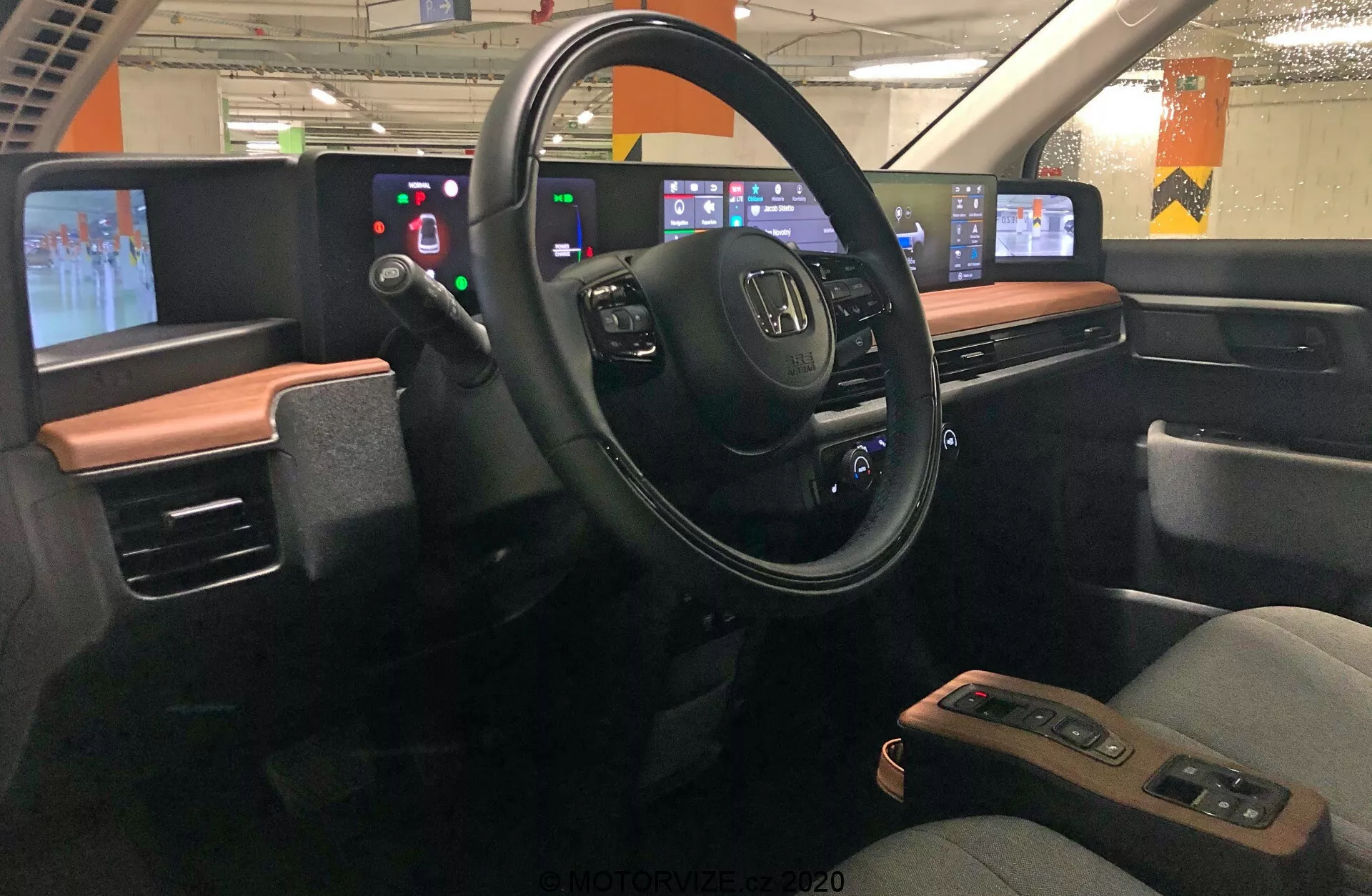 Interiörvy av 2020 Honda e, fokuserad på förarområdet sett från mitten av fordonet. Instrumentpanelen visar en modern design med ett tvåfärgat färgschema. Ratten, med Honda-logotypen och flera kontrollknappar, är i mitten, omgiven av en digital displaypanel istället för analoga mätare. Till höger finns den digitala mittkonsolen med pekskärmar för ljud, navigering, klimatkontroll och andra inställningar. Designen betonar teknikintegration och minimerar fysiska knappar till förmån för pekskärmsgränssnitt. Bilen verkar stå still i ett parkeringshus, med lysrörsbelysning och betongpelare synliga genom fönstren.