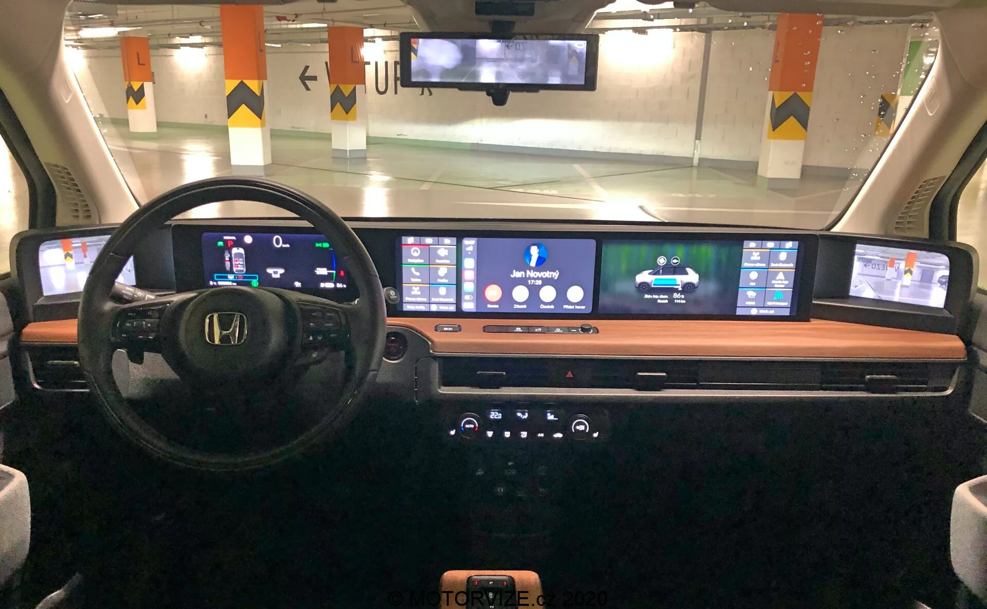 Interiörvy av instrumentpanelen i Honda e 2020 från förarplatsens perspektiv, med digital instrumentpanel, multifunktionell ratt, expanderande sammanhängande digital skärm för navigering och media, elegant mittkonsol med knappar och reglage och en trådlös telefonladdningsplatta, framhäver modern bilteknik och design.
