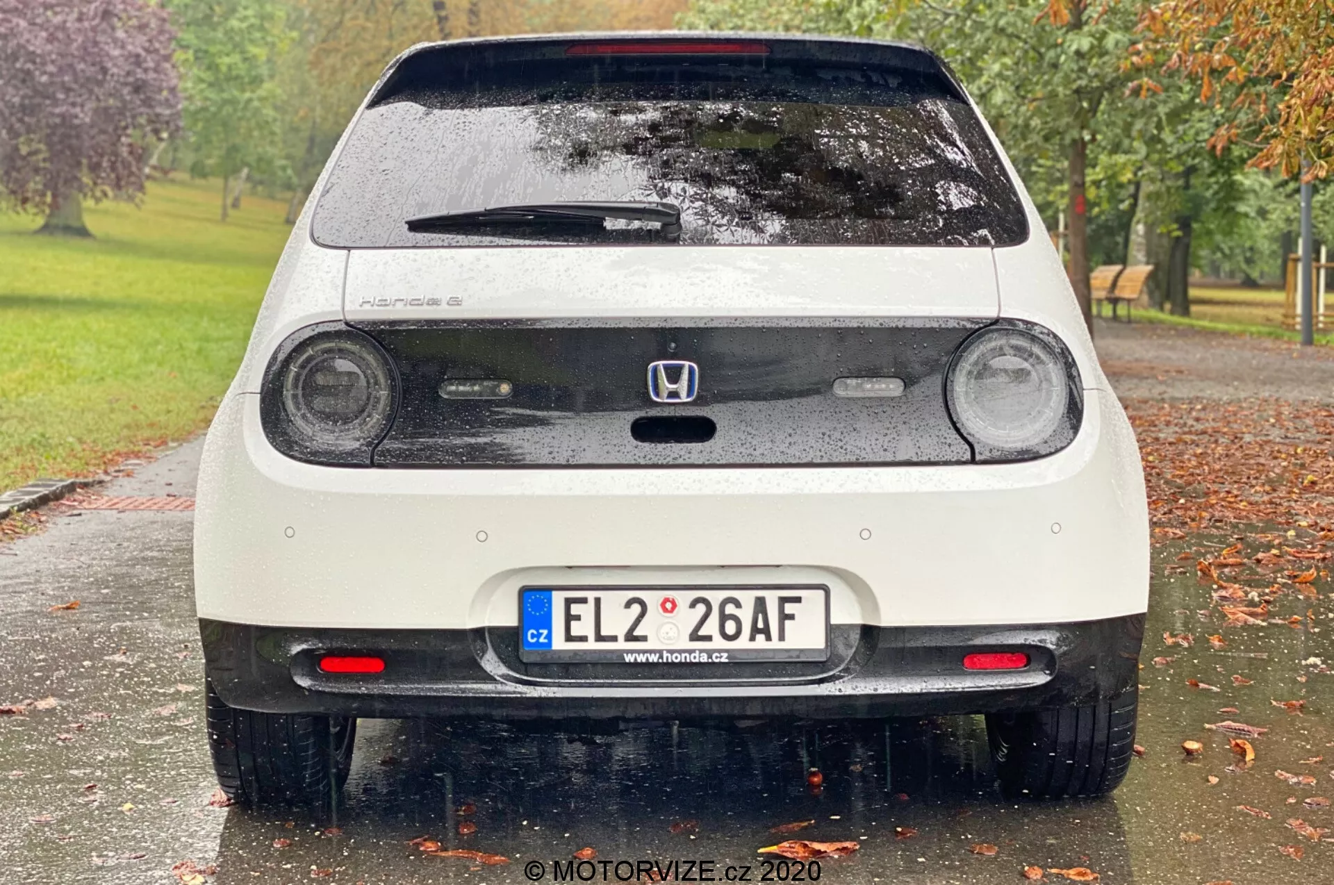 Bakifrån på en vit och blöt Honda e 2020, som visar cirkulära LED-bakljus, tredje bromsljus, Honda-emblem, baklucka med vindrutetorkare, europeisk registreringsskylt, parkeringssensorer, bakre dimljus, bakspoiler och reflekterande element på stötfångaren. Bilden fångar fina detaljer som vattendroppar på fordonet, med en suddig parkmiljö med träd och gräs i bakgrunden.
