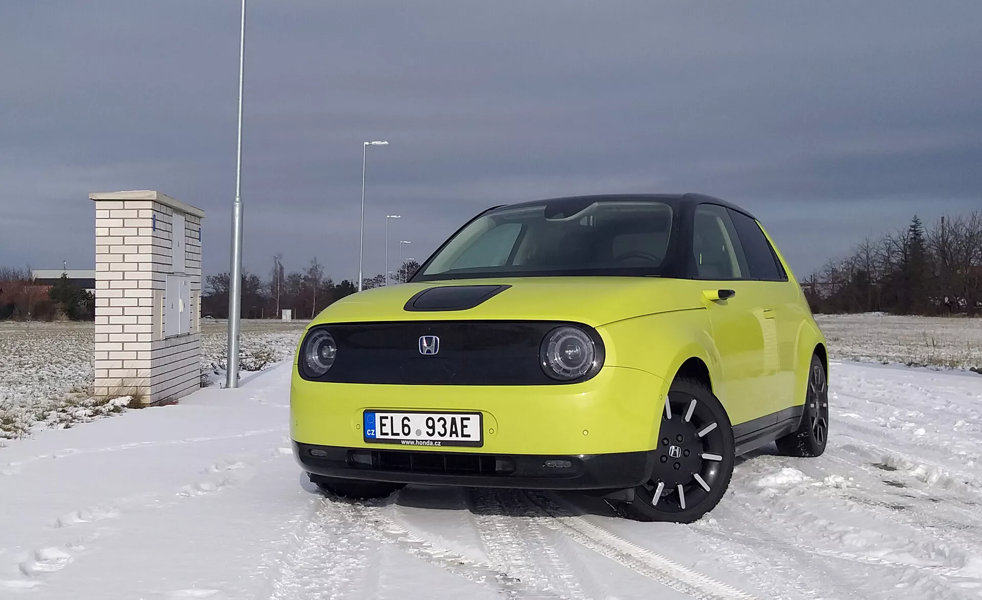 Honda e 2020 i klar gult på snötäckt yta med molnig himmel, med LED-strålkastare, elbil teknologi med avancerade förarassistanssystem, svarta legeringshjul och vinterdäck.