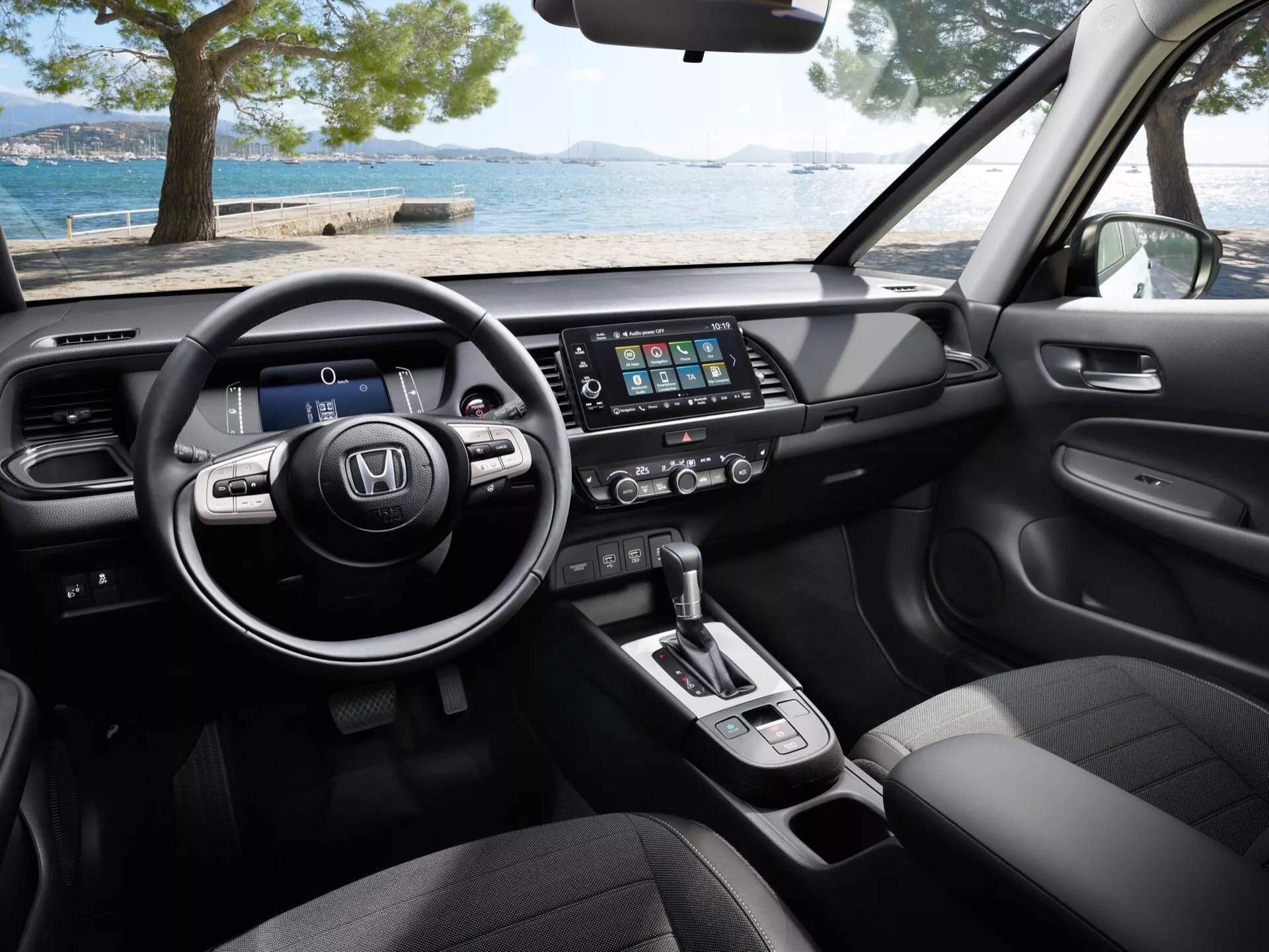 Närbild av interiören i Honda Jazz 2023 (5:e generationen, ansiktslyft), med fokus på dess moderna läderklädda multifunktionsratt, digitala instrumentbräda, pekskärmsbaserade underhållningssystem, klimatkontroll, automatisk växelspak, tygsäten och premiummaterial från främre passagerarens perspektiv.