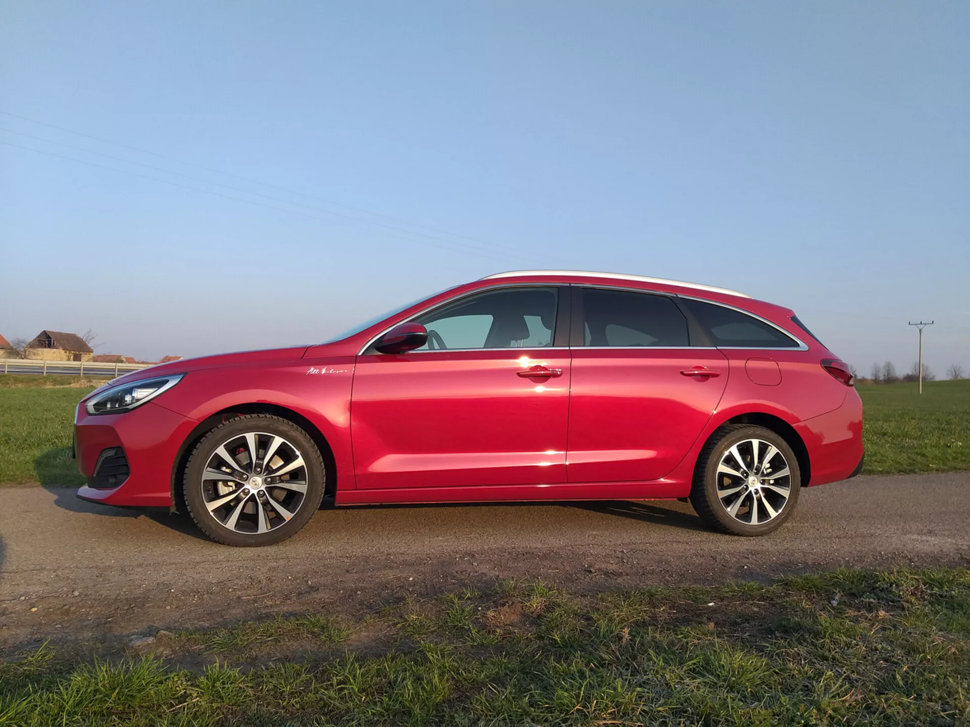 Röd Hyundai i30 Fastback från 2017, med en modern aerodynamisk profil med en sluttande taklinje, flerspakiga legeringshjul, sidospeglar med integrerade blinkers, fönster med kromram och vägklara däck, parkerad på en gräsmatta under en klar himmel.