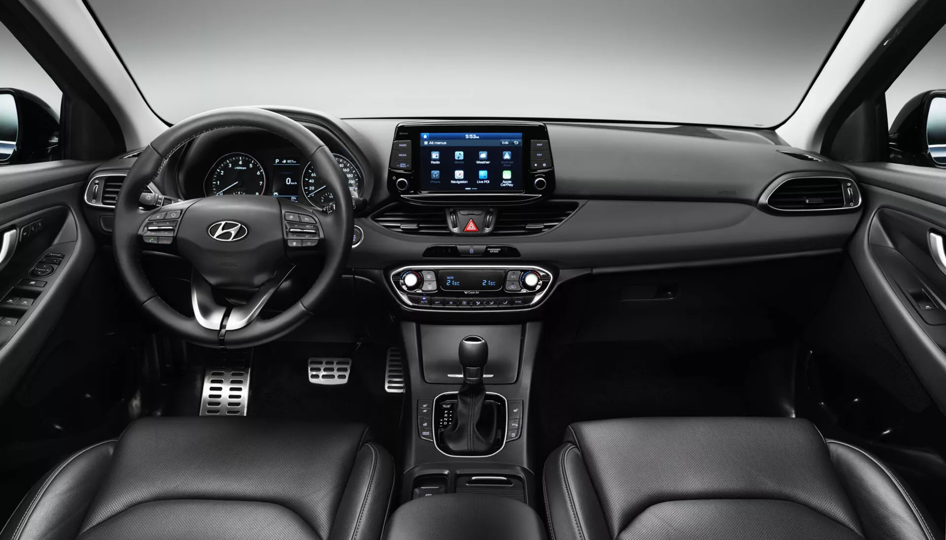Interiör av Hyundai i30 2017 Fastback som visar främre instrumentbrädan från ett baksätesperspektiv, med ratten placerad i mitten till vänster med kontrollknappar och Hyundai-logotyp, en digital instrumentpanel bakom, ett stort pekskärmsinfotainmentsystem med anslutningsappar, dubbelzons klimatkontroll, automatisk växelspak, eventuella kontroller för sätesuppvärmning/kylning och säten klädda i läder.