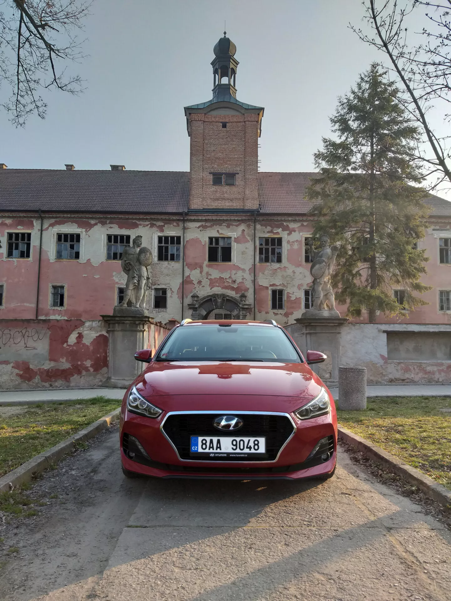 2017 års Hyundai i30 Fastback i rött sett framifrån med full vy över LED-strålkastare och skulpterad grilldesign, positionerad på en grusgång med en historisk byggnad i bakgrunden.