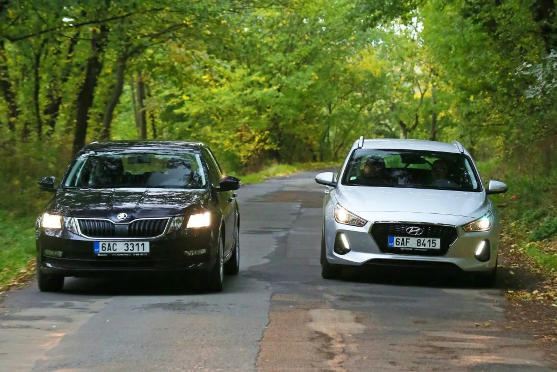 Två bilar står inför varandra på en trädbeprydd landsväg i tidig höst: en mörkfärgad Skoda med rektangulär grill och standardstrålkastare till vänster, och en ljusfärgad Hyundai i30-N med kurvlinjig front, moderna projektionsstrålkastare och integrerade dimljus till höger som illustrerar en jämförelse mellan traditionella och moderna bil designelement.