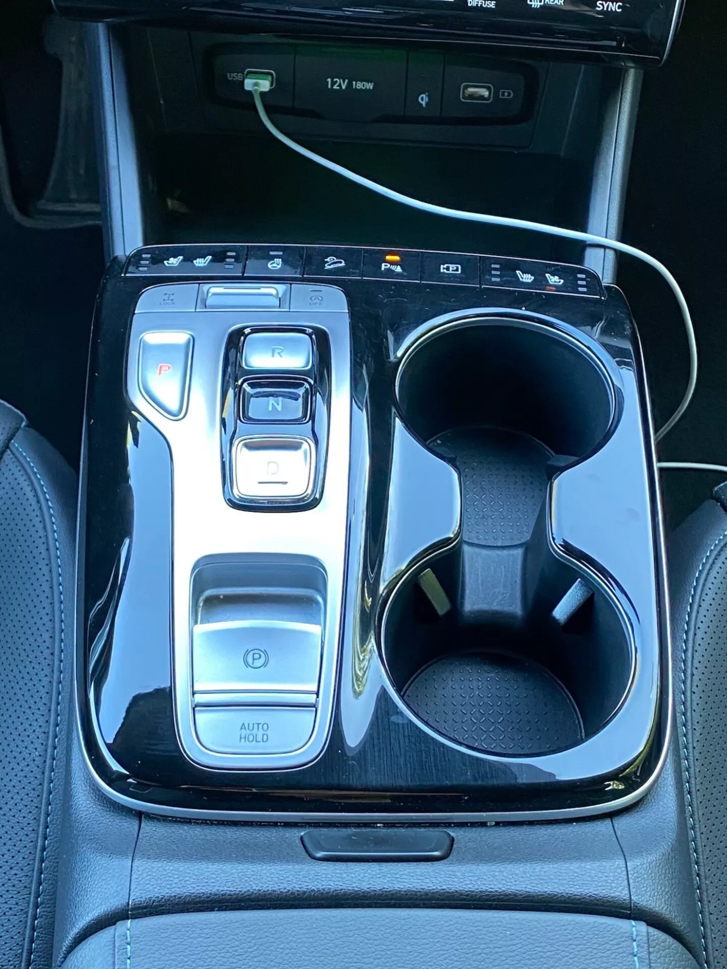 Bilden visar mittkonsolen i en Hyundai Tucson 2020, med en elektronisk växelväljare i läge 'N' omgivet av en blank svart panel. Till vänster finns kontroller inklusive en strömbrytare, körlägesväljare och funktionen 'AUTO HOLD'. Till höger finns dubbla mugghållare, varav en med en vit laddningskabel. I övre delen syns en del av en infotainmentskärm med USB-port indikationer, vilket framhäver bilens teknik och lyx.