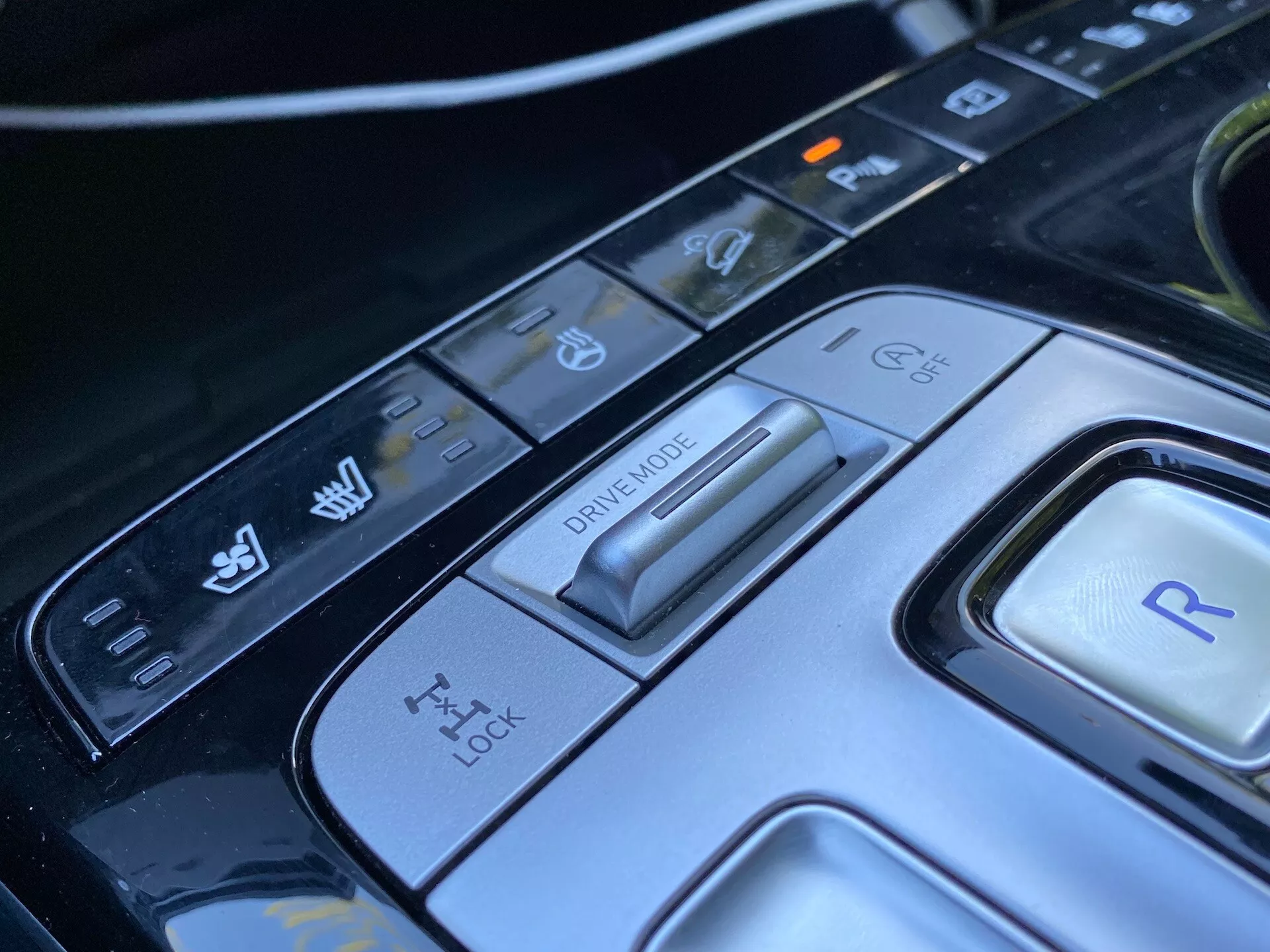 Närbild på Hyundai Tucson 2020:s mittkonsol, med fokus på körlägesväljaren och olika kontrollknappar. Bilden visar en avancerad körlägesväljare för olika körförhållanden, en kontrollpanel för traktionskontroll, en knapp för backväxel och ytterligare kontroller för fordonets stabilitet och parkeringsassistansfunktioner. Den eleganta designen och högteknologiska materialen framhäver det sofistikerade interiöret i denna lyxbil.