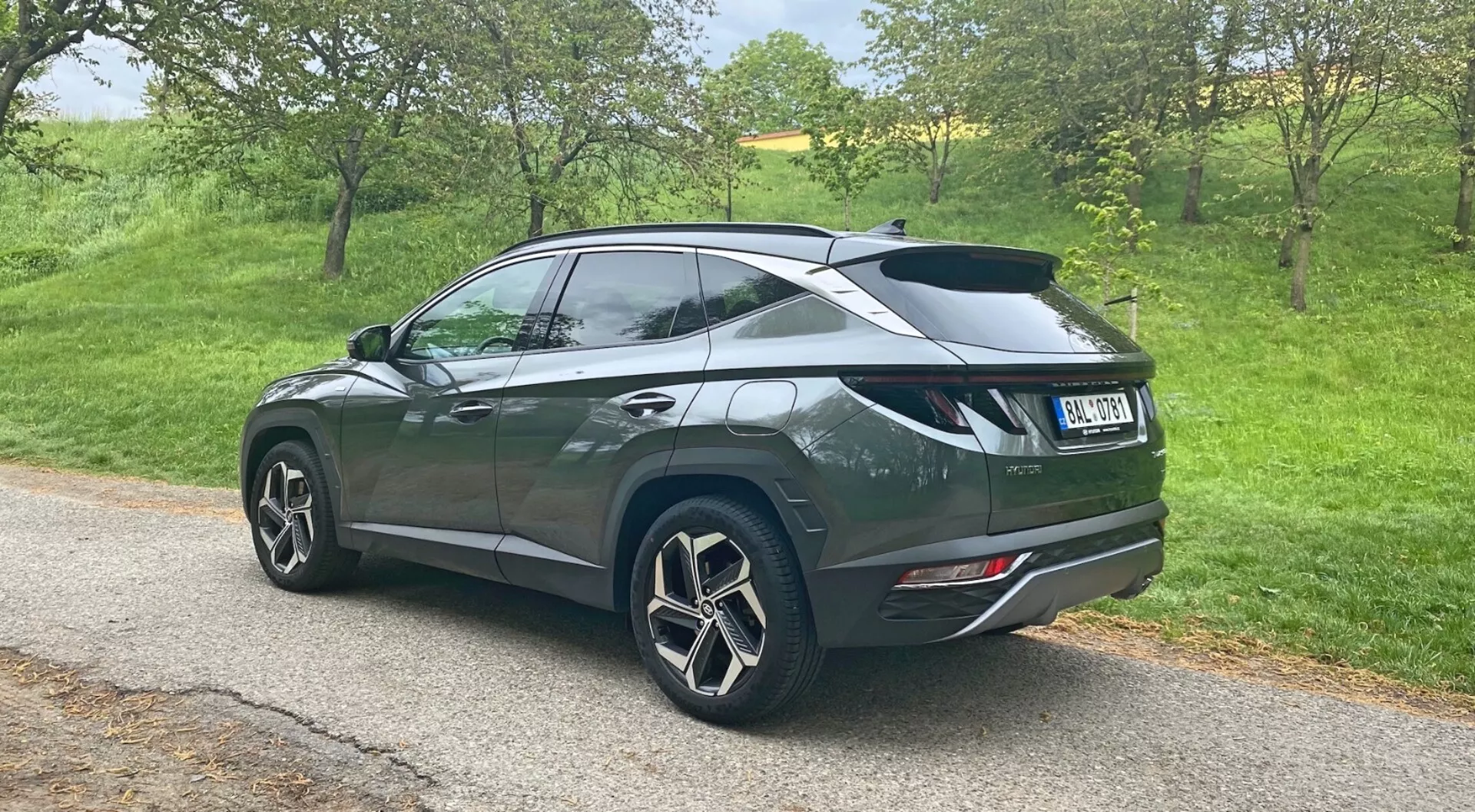 En tredjedels bakifrån perspektiv av en 2020 Hyundai Tucson 1.6 T-GDi Pure SUV parkerad på en smal väg i lantlig miljö, som visar stora flerspoked aluminiumfälgar, mörk metallisk lack med en sluttande taklinje, bakspoiler, privatglas på bakrutorna, moderna bakljus och dubbla avgasrör integrerade i den bakre stötfångaren.