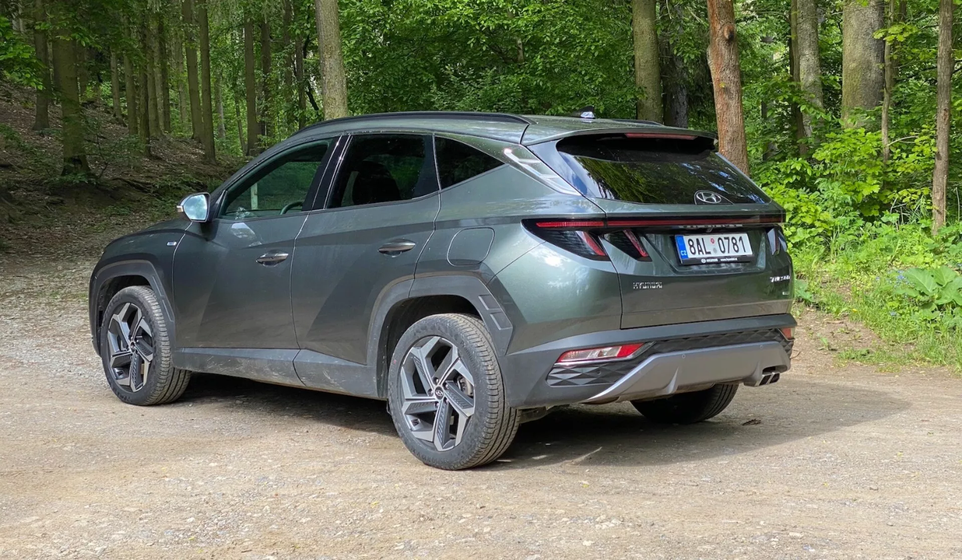 En modern SUV parkerad på en grusväg i ett skogsområde, fotad från en bakre trekvartsvy från en lätt höjd. Fordonet har en elegant, modern design med skarpa linjer, förlängda baklyktor som sträcker sig från baksidan till sidorna, en synlig spoiler och reflektorer på den bakre stötfångaren, vilket antyder avancerade säkerhetsfunktioner. Det metalliska gråa fordonet har flerekrade aluminiumfälgar och vägdäck, med en glänsande yta som indikerar att det är nytt eller väl underhållet. Runt bilen finns en grön, frodig skog med packad jord och lösgrus på marken.