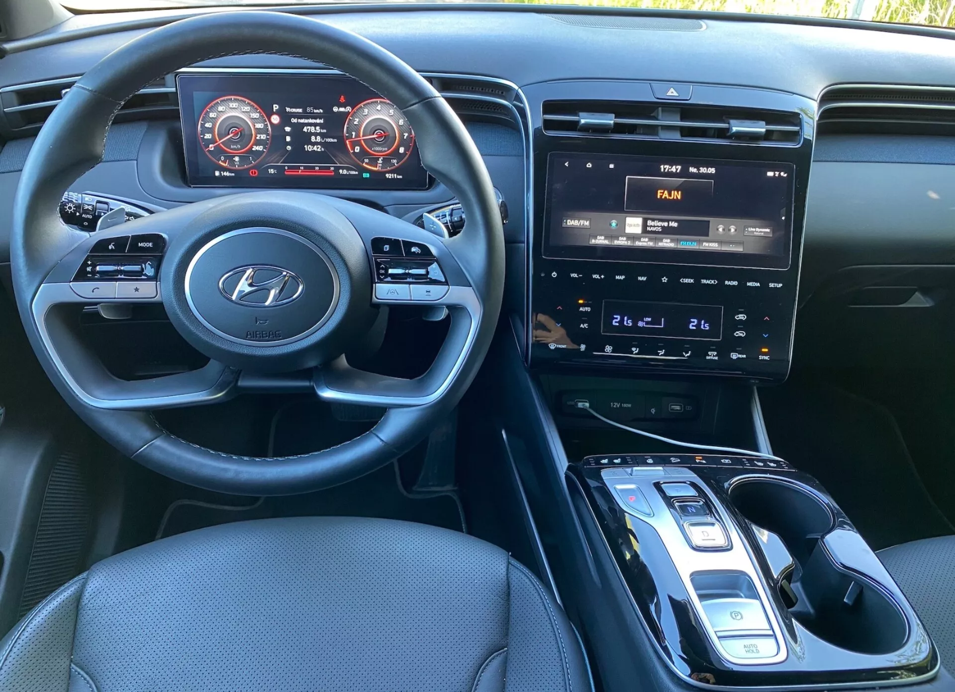 Inre vy av Hyundai Tucson 2020 från förarsätet, som visar ratten med Hyundai-logotypen och monterade kontroller för ljud, telefon och farthållarinställningar. Instrumentpanelen innehåller en digital instrumentbräda med rödmarkerade hastighets- och varvräknare. Mittkonsolen rymmer ett infotainmentsystem med pekskärm som visar luftkonditioneringskontroller, fysiska knappar och vred för klimatkontroll, och en automatisk växelspak med elektronisk växelväljare. En potentiell trådlös laddningsplatta är synlig, tillsammans med mörk tygklädsel och en väl upplyst hytt.