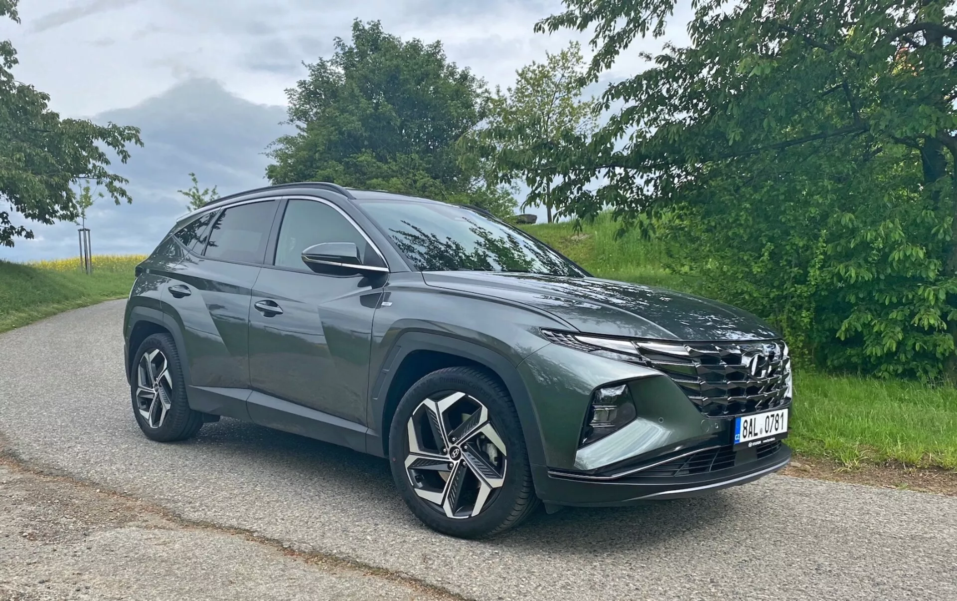 Bilden visar en modern Hyundai Tucson 1.6 T-GDi Pure SUV parkerad på en asfalterad väg med naturligt landskap i bakgrunden. Fotograferad från en främre trekvartsvinkel syns bilens framträdande grill, eleganta strålkastare, främre stötfångare med dimljus eller luftintag, stora lättmetallfälgar med en flerradie design och sidospeglar. SUV:ens siluett har en sluttande taklinje bakåt, vilket indikerar en sportig, aerodynamisk design. Fotot har tagits i mjuka dagsljusförhållanden, vilket framhäver fordonets färg och designdetaljer utan hårda skuggor.