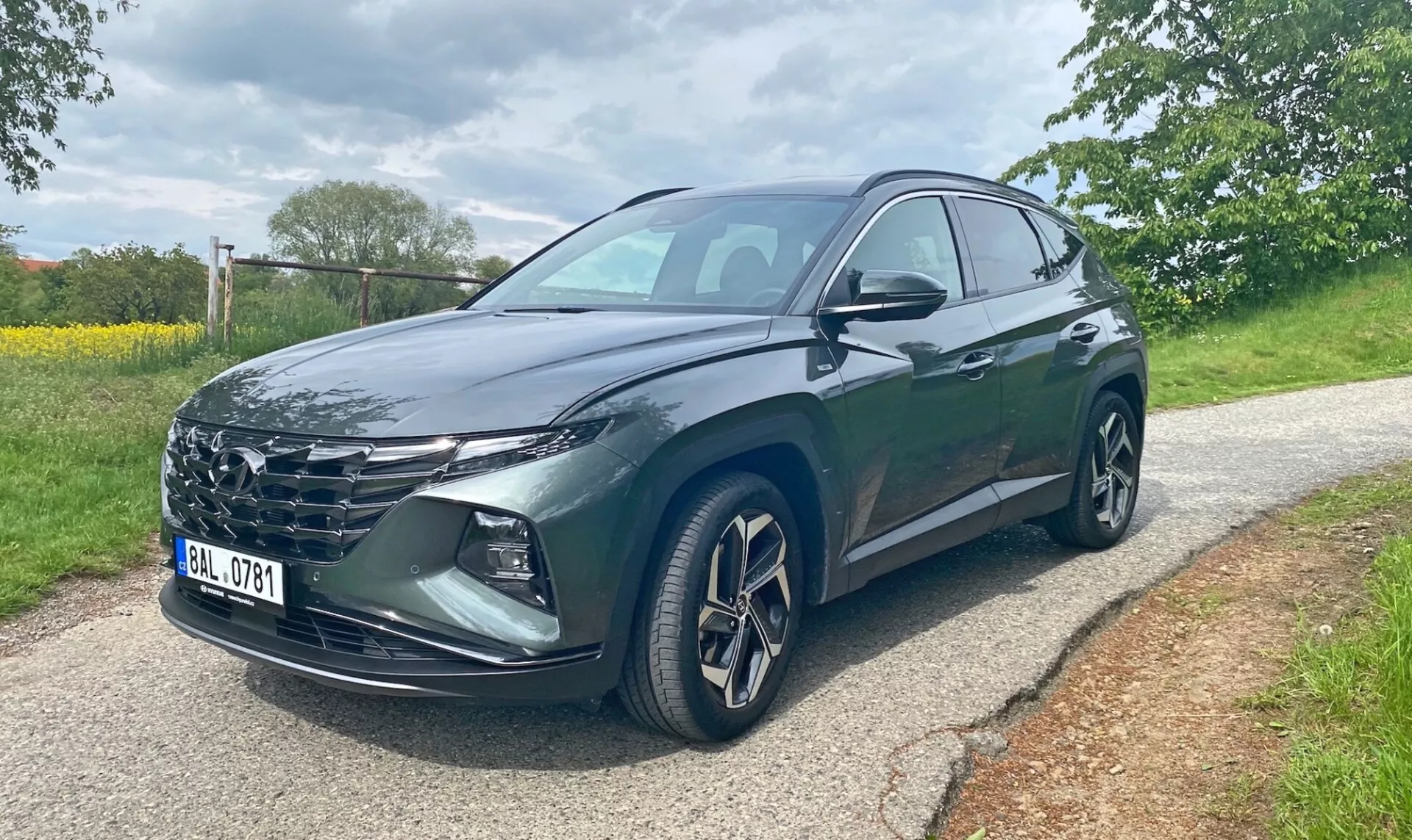 Hyundai Tucson 2020 1.6 T-GDI Pure SUV, sedd från en trekvarts vinkel framifrån på en asfalterad väg med träd och gräs i bakgrunden. Bilden visar fordonets grill, skarpa LED-strålkastare, lättmetallfälgar, sidospeglar med integrerade blinkers och en mörk metalliclack. Förarens fram- och bakdörrar är synliga. Fotot togs under dagtid i mulet ljus.