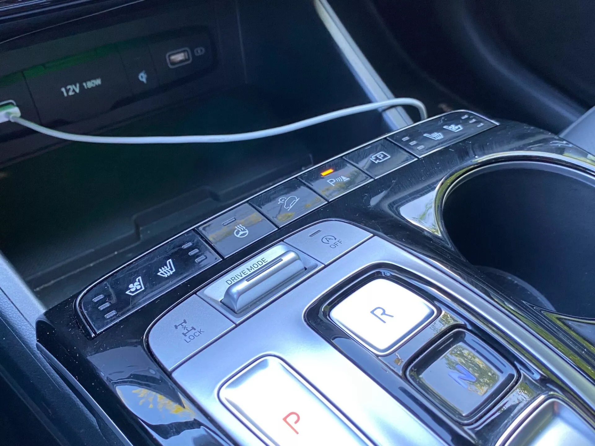 Närbild av den centrala konsolen och växelområdet i en Hyundai Tucson 2020 1.6 T-GDi Pure, med modern design med knappar för parkering, backning, neutral, körning och broms/regenerativa lägen, tillsammans med ytterligare knappar för auto start/stop, parkeringssensorer/automatisk parkering och körfältsassistans-/avvikelsevarning. Synligt multimediasystem, kopphållare och 12V-uttag som indikerar komfort, bekvämlighet och avancerade förarassistanssystem (ADAS). Konsolen har en elegant design med svart glansigt material och metallaccent, vilket antyder en lyxig känsla.