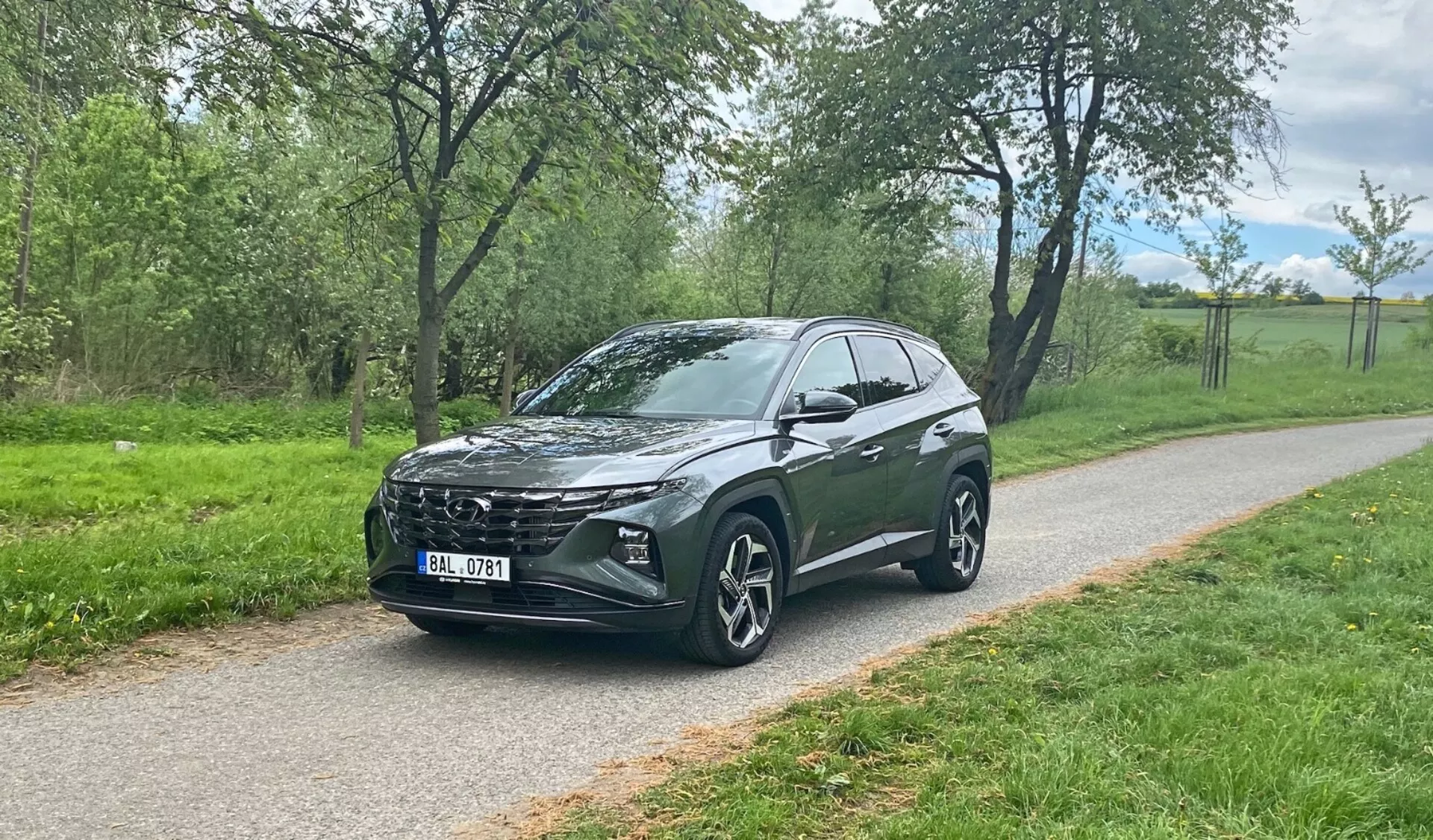 Trekvarts frontal vy av en Hyundai Tucson 2020 crossover SUV parkerad på en asfalterad väg med grönska i bakgrunden, som visar fordonets distinkta frontgrill, eleganta LED-strålkastare, stora multifälgar i aluminium, sluttande taklinje, hög markfrigång, integrerade sidospeglar, tonade fönster och stiliga karosskonturer.