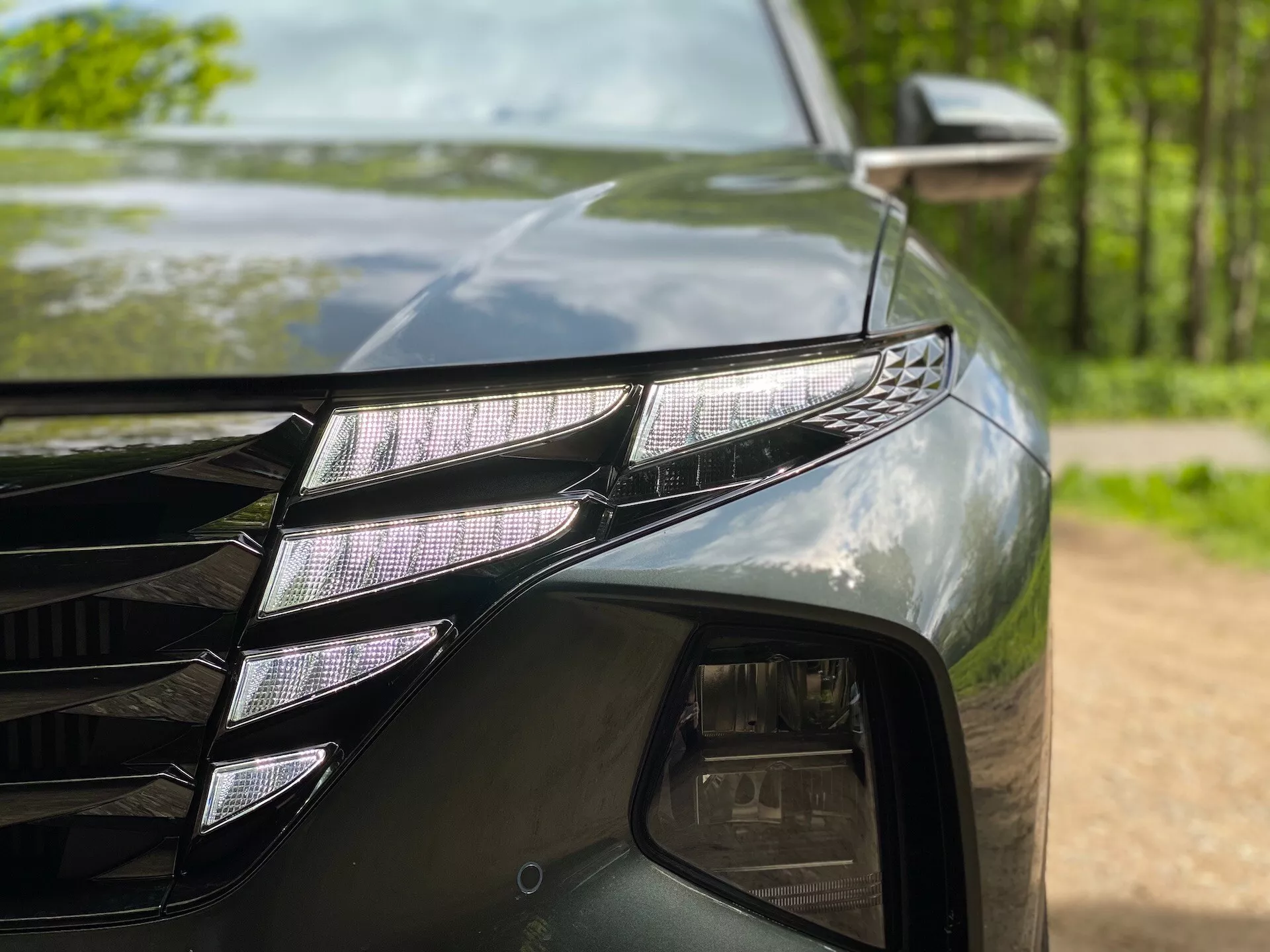 Närbild av den vänstra främre delen av en Hyundai Tucson 2020 (Hyundai-Tucson-1-6-T-GDi-Pure), som visar en LED-strålkastarenhet med individuella LED-projektorer och en distinkt LED DRL-remsa. Den mörka, glansiga färgen speglar omgivande träd. En del av grillen med kantiga linjer och sidospegeln med en blinkersindikator är också synliga.