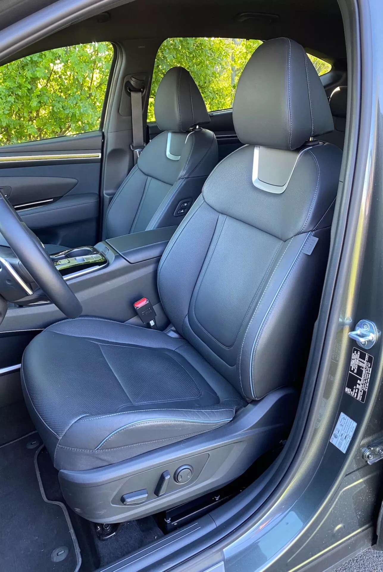Interiören av 2020 Hyundai Tucson 1.6 T-GDI Pure som visar framsätet för passageraren med svart läderklädsel, detaljerad sömnad och elektriska justeringskontroller. Synlig mittkonsol med mugghållare, dörrpanel med matchande material, handtag, förvaringsfack och högtalargaller som antyder ett avancerat ljudsystem. Delvis synlig instrumentpanel och handsfack. Mattliknande golvmatta och lätt solljusreflektion på dörrpanelen.