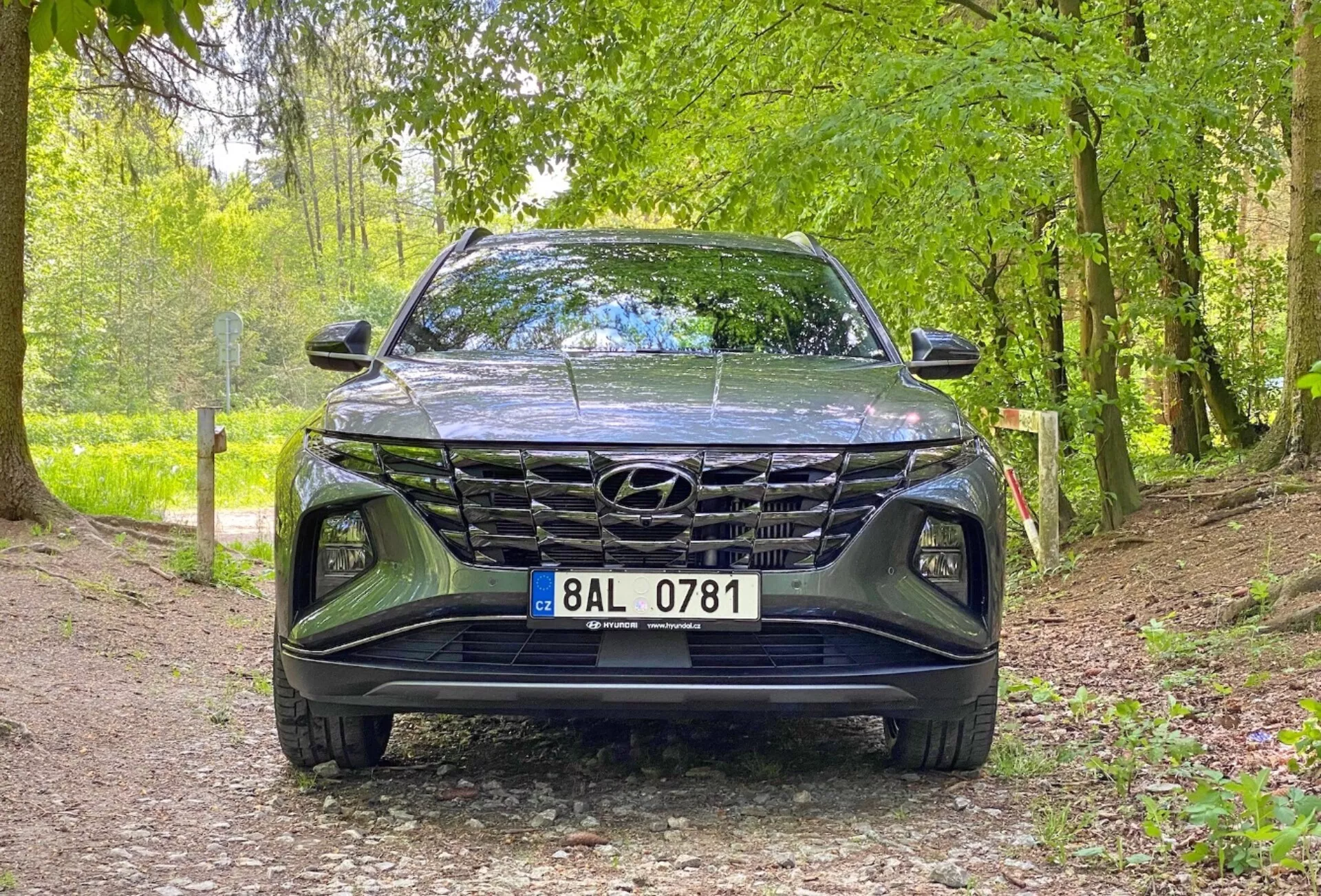 Frontal vy av 2020 Hyundai Tucson 1.6 T-GDi Pure-fordon, som visar en låg vinkelbild fokuserad på grillen, främre stötfångaren och huvområdet. Bilen är parkerad på en grusväg i en naturlig miljö, vilket antyder dess lämplighet för utomhusmiljöer. Nyckelfunktioner inkluderar en framträdande grilldesign, eleganta LED-strålkastare och en synlig Hyundai-emblem.